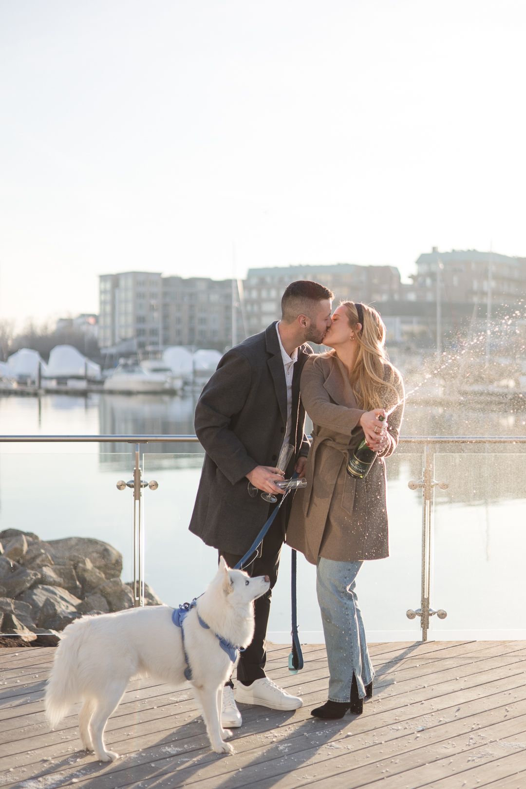A romantic couple shares a kiss on a marina dock with their white dog on a sunny afternoon.