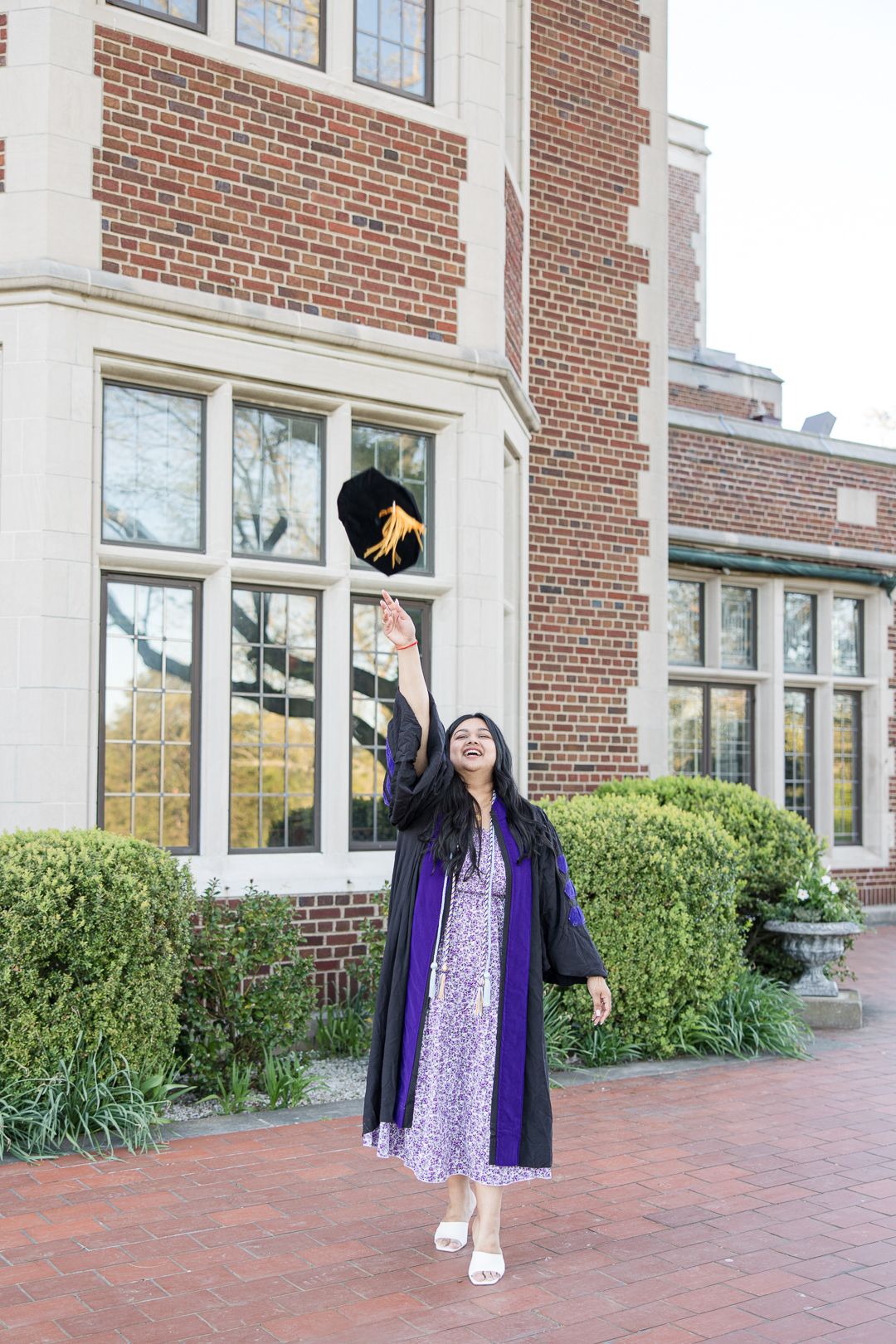 Graduate in purple dress and regalia throws cap in celebration outside brick building.