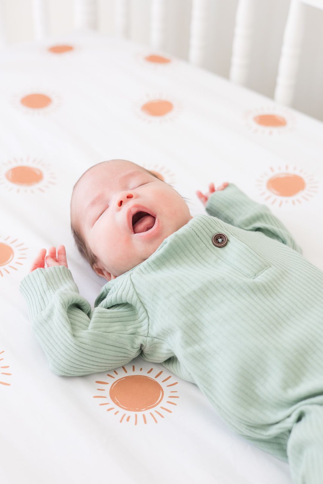 A newborn baby in mint green clothing yawns while lying on a sun-patterned crib sheet.