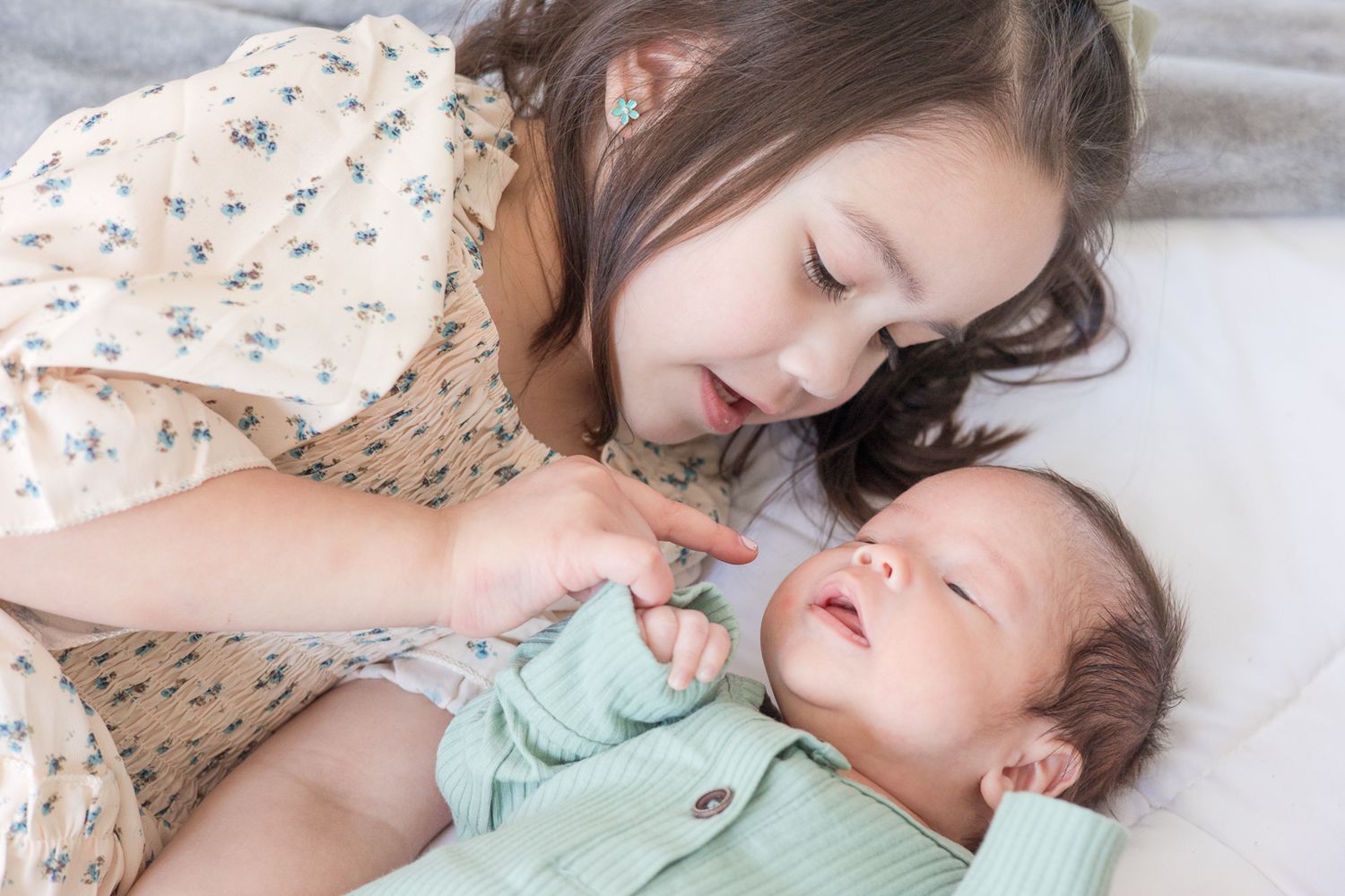 Two siblings share a sweet moment with a baby wearing mint green on white bedding.