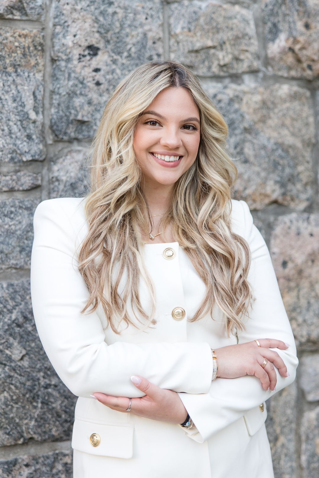 A woman in a white blazer with gold buttons poses against a stone wall with wavy blonde hair.