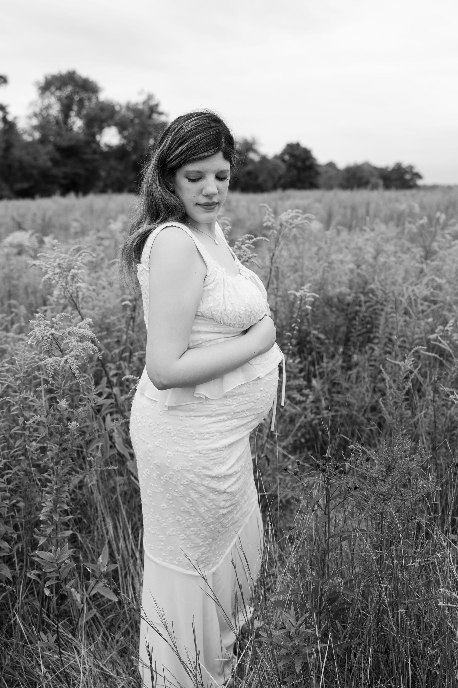 Expectant mother in a white dress stands peacefully in a wildflower meadow during a black and white maternity photoshoot.