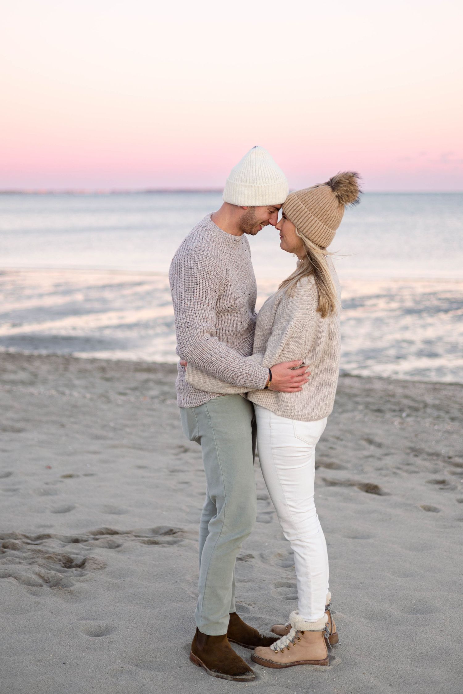 Couple in winter clothing embrace on beach during pink sunset.