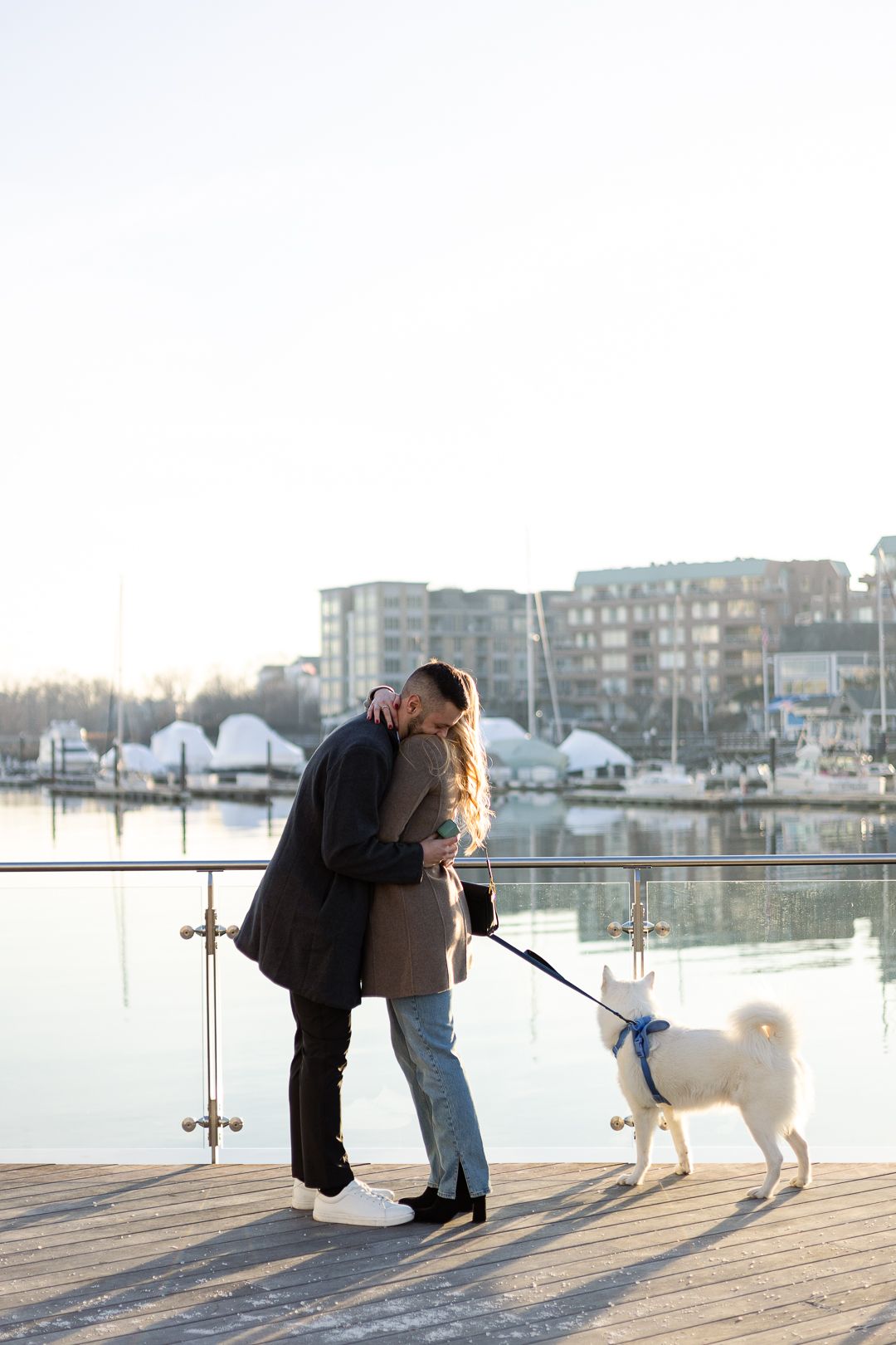 Couple embracing on marina boardwalk with white dog on leash during golden hour.