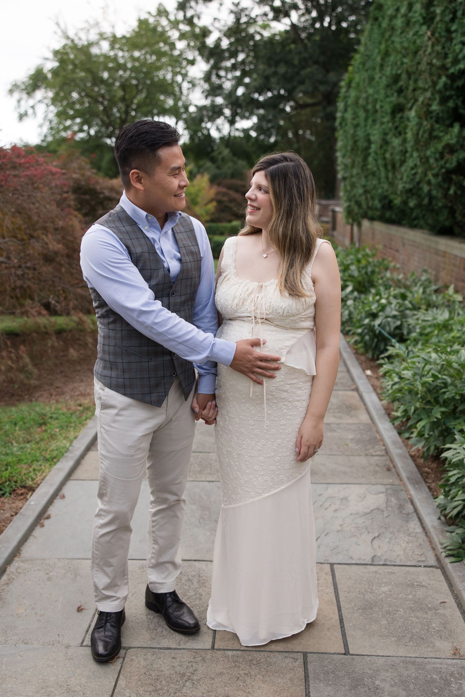 Expectant couple shares tender moment on stone pathway surrounded by garden foliage.