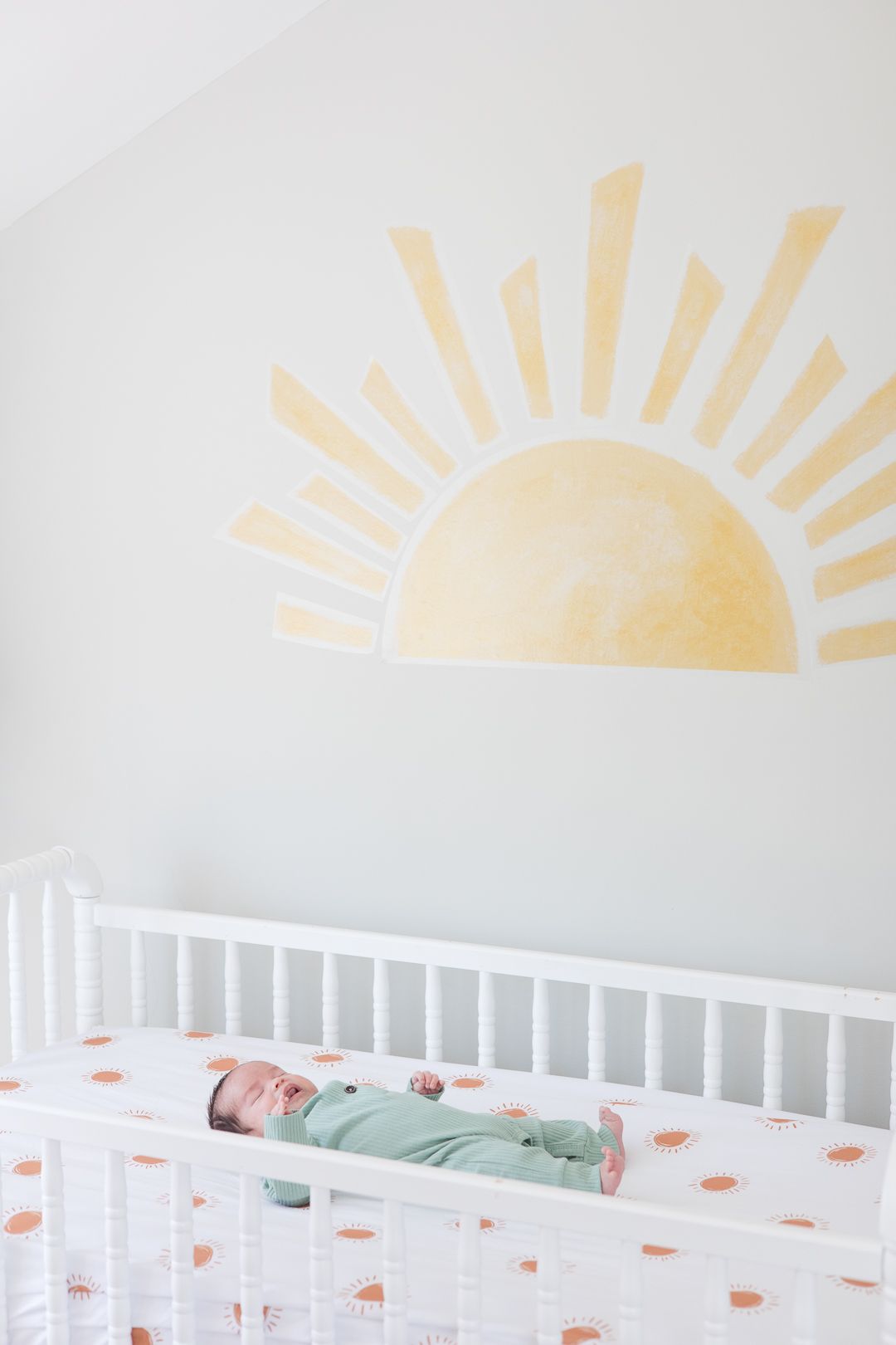 A sleeping newborn in mint green sleepwear lies in a white crib beneath a yellow sun wall decal.