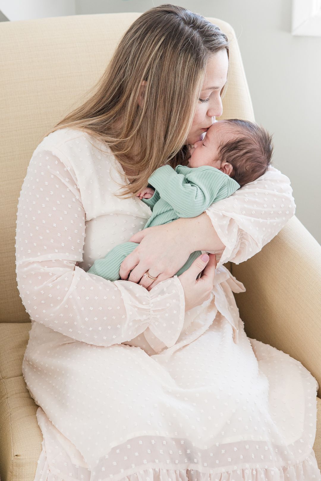 A woman in a white dress tenderly holds and kisses a sleeping newborn baby in mint green sleepwear.