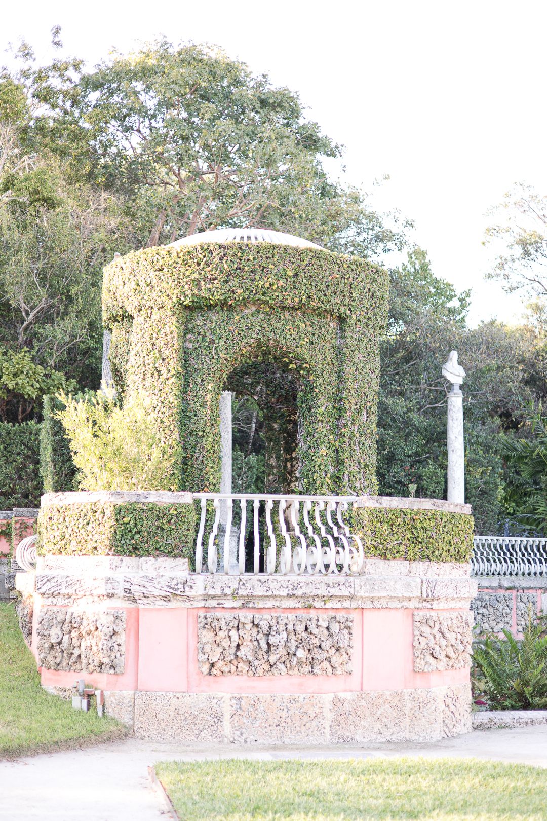 Pink stone gazebo covered in climbing vines stands elegantly in a formal garden.
