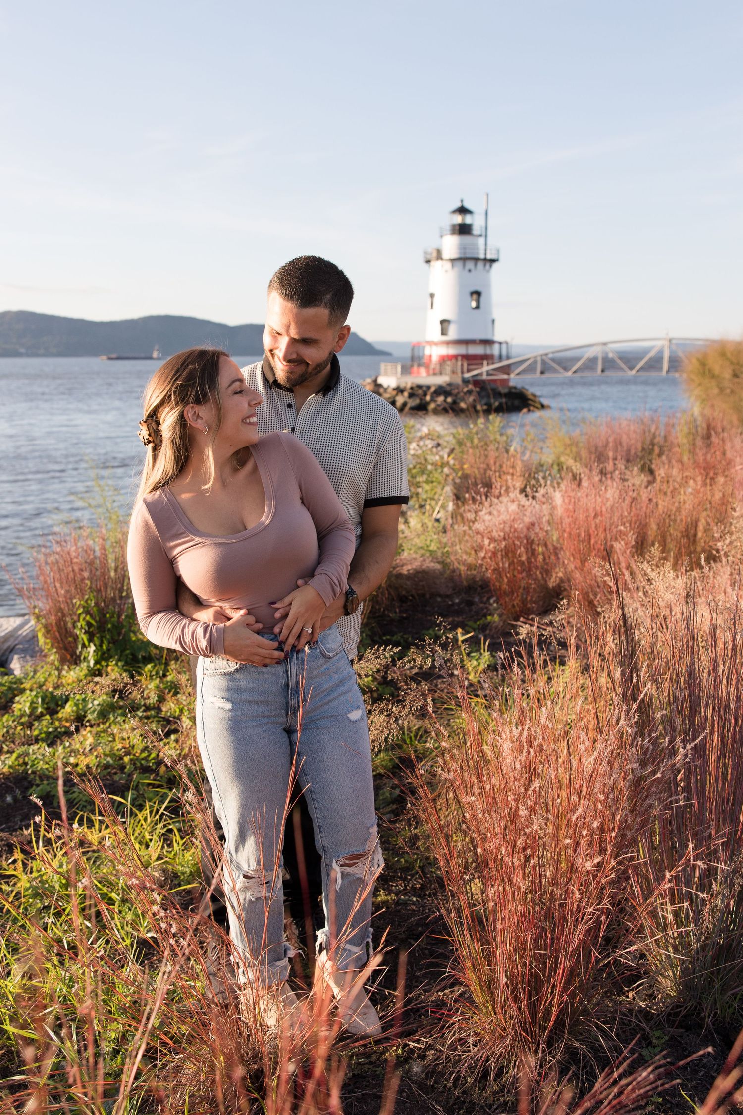 Romantic couple embracing by waterfront lighthouse with ornamental grasses at sunset.