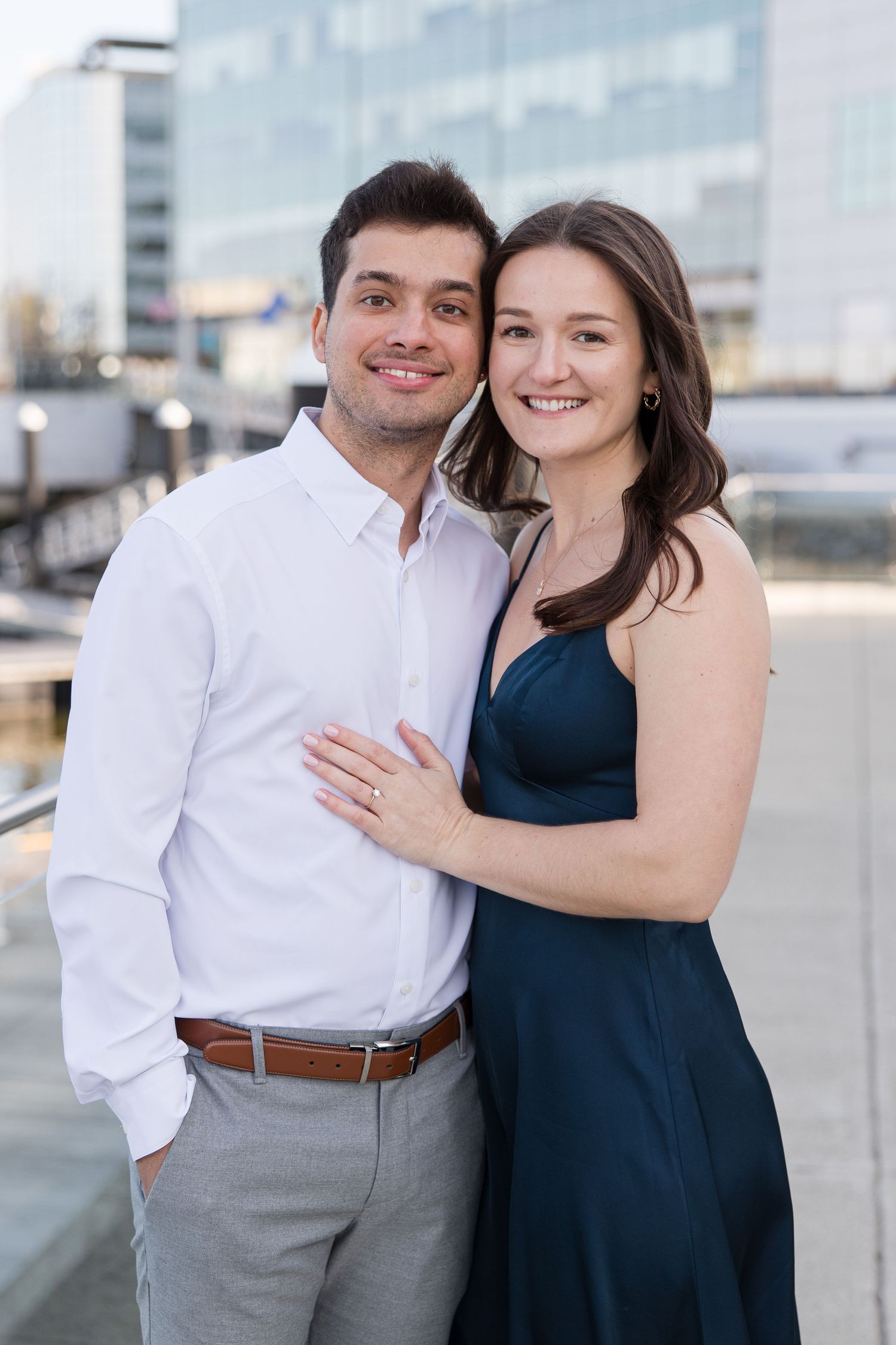 A couple poses together on a waterfront with modern buildings in the background.