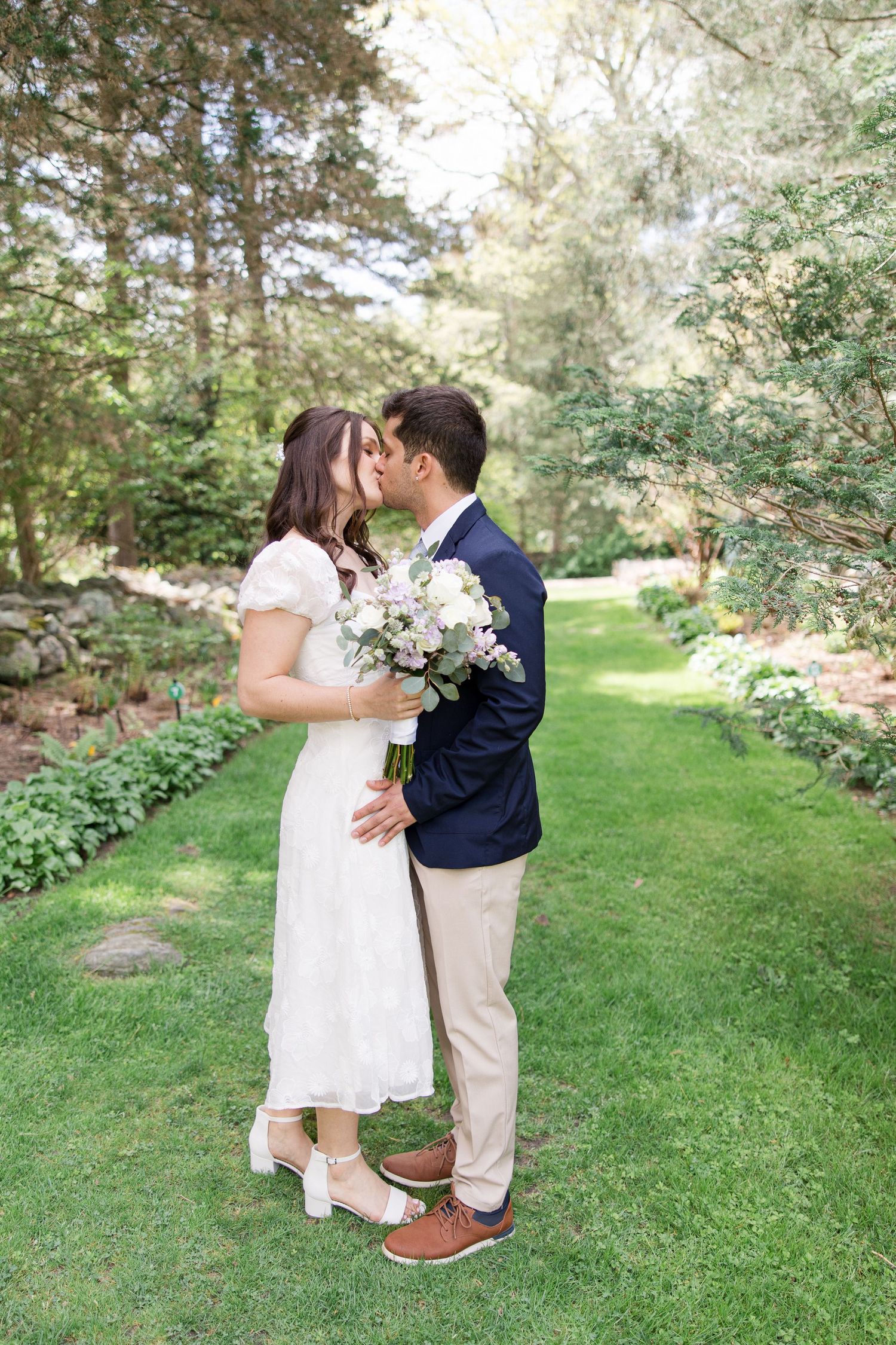 A romantic embrace between newlyweds on a manicured garden lawn surrounded by evergreens.