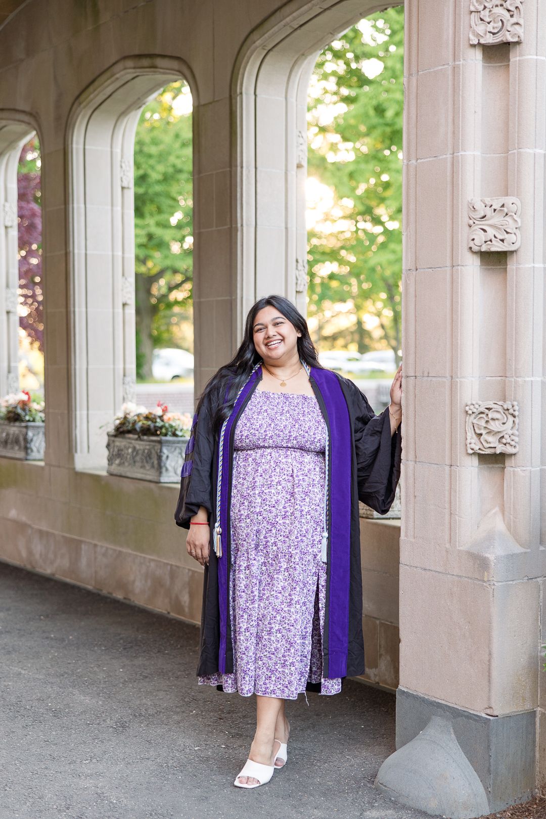 Graduate in purple floral dress and academic regalia poses under stone archway.