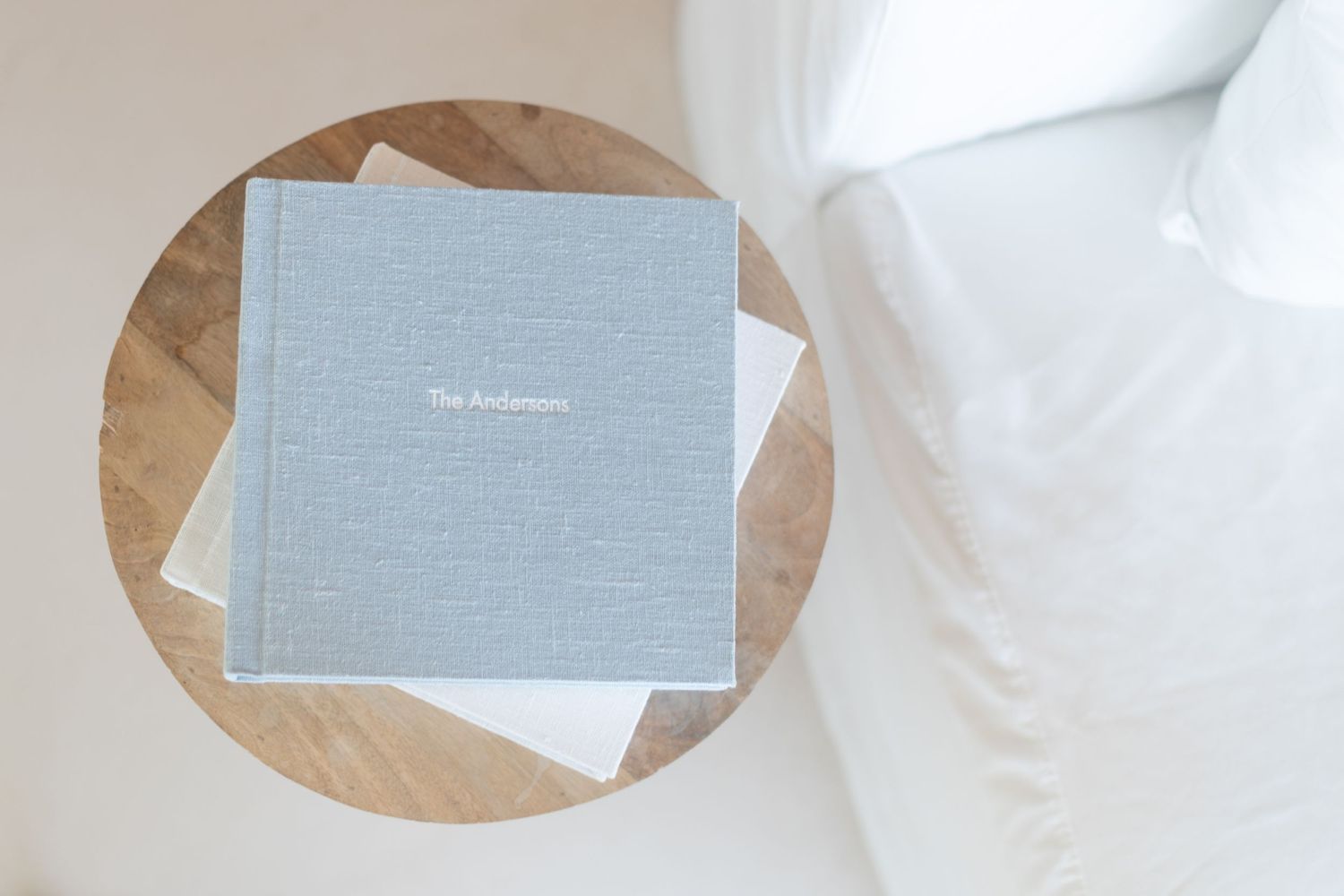 A light blue photo album book rests on a round wooden side table next to white bedding.