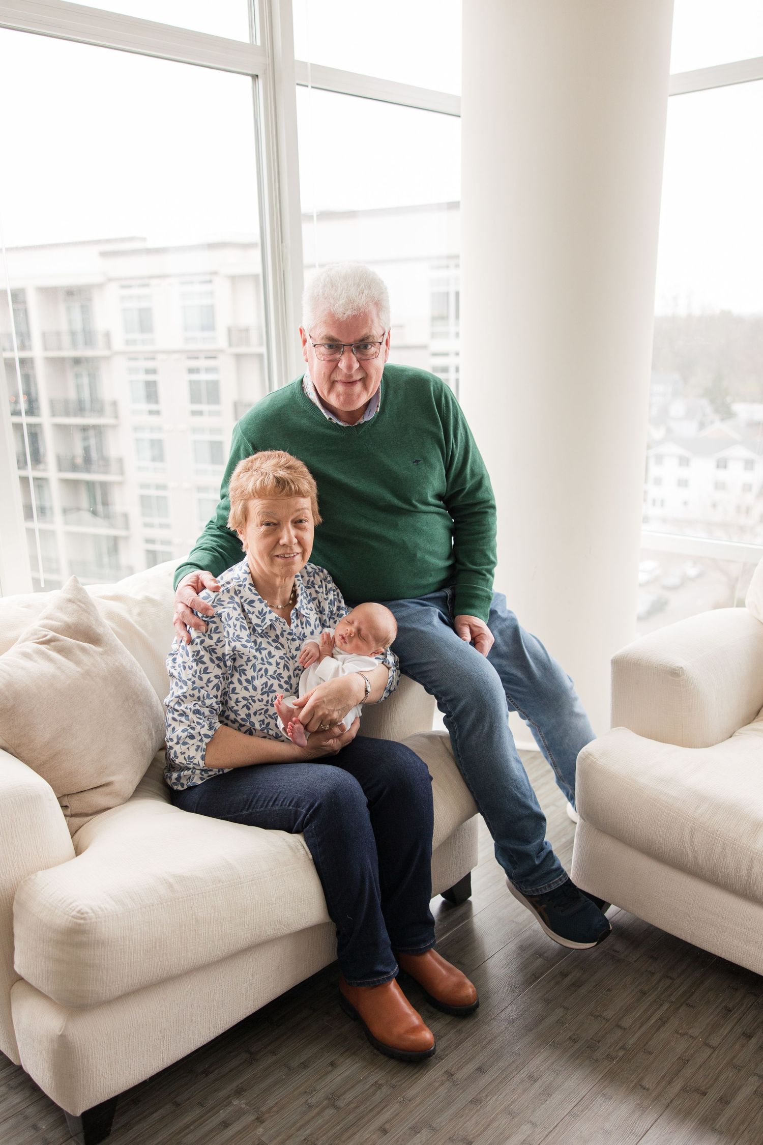 A senior couple sits together on a white couch by large windows in a bright, modern living room.