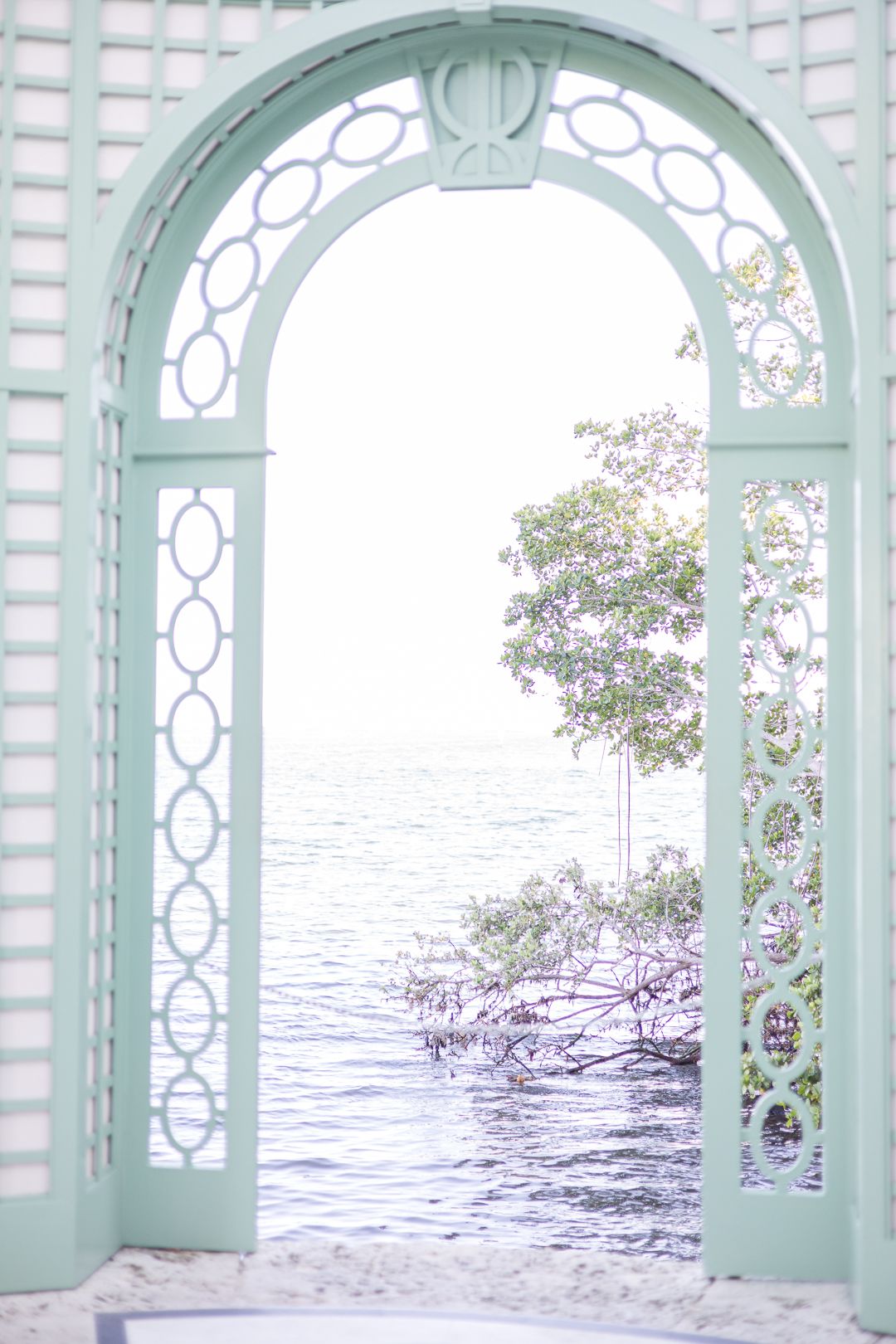 A mint green latticed archway frames a tranquil ocean view with mangroves in soft pastel tones.