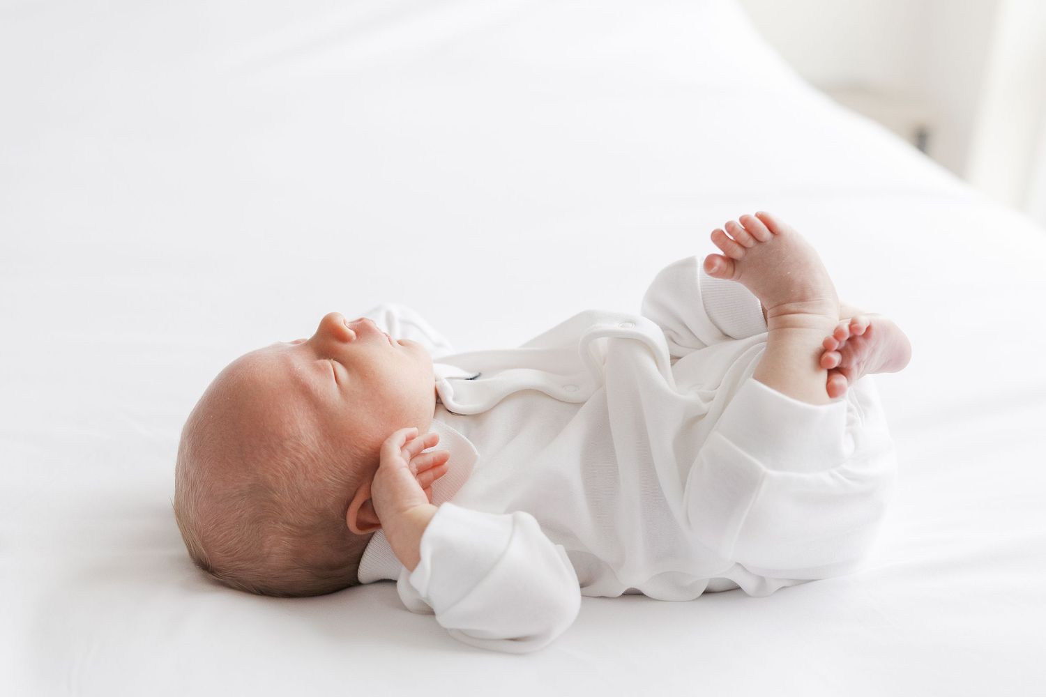A peaceful baby in white clothing lies on a white blanket, stretching their tiny arms and legs.