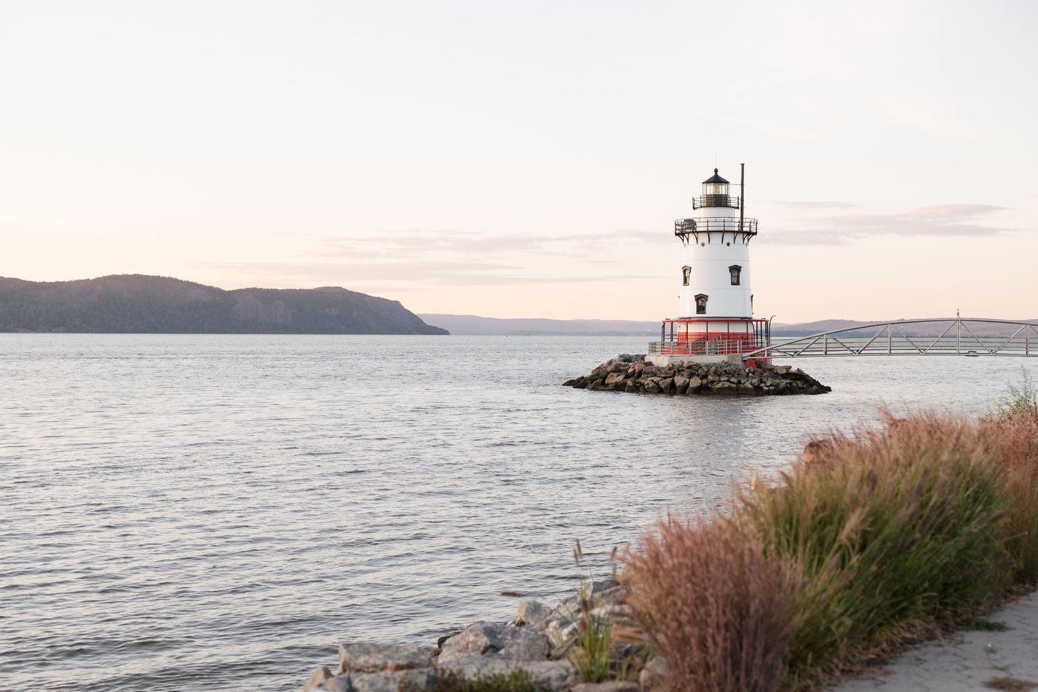 Scenic lighthouse on rocky peninsula with calm waters and mountains in distance during sunset.