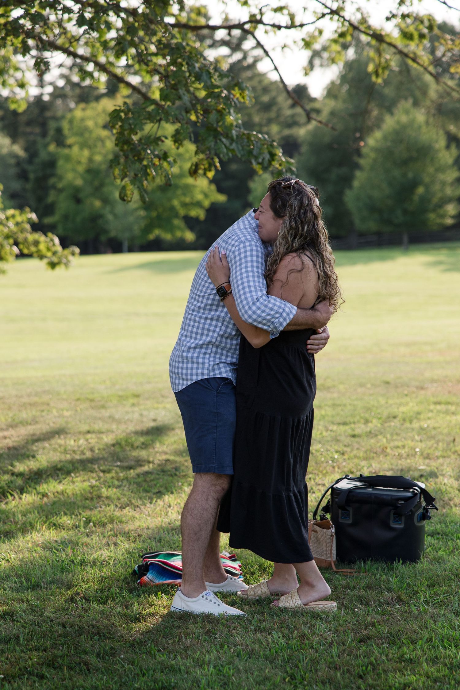 A tender embrace between two people in a sunlit park with a tree in the background.
