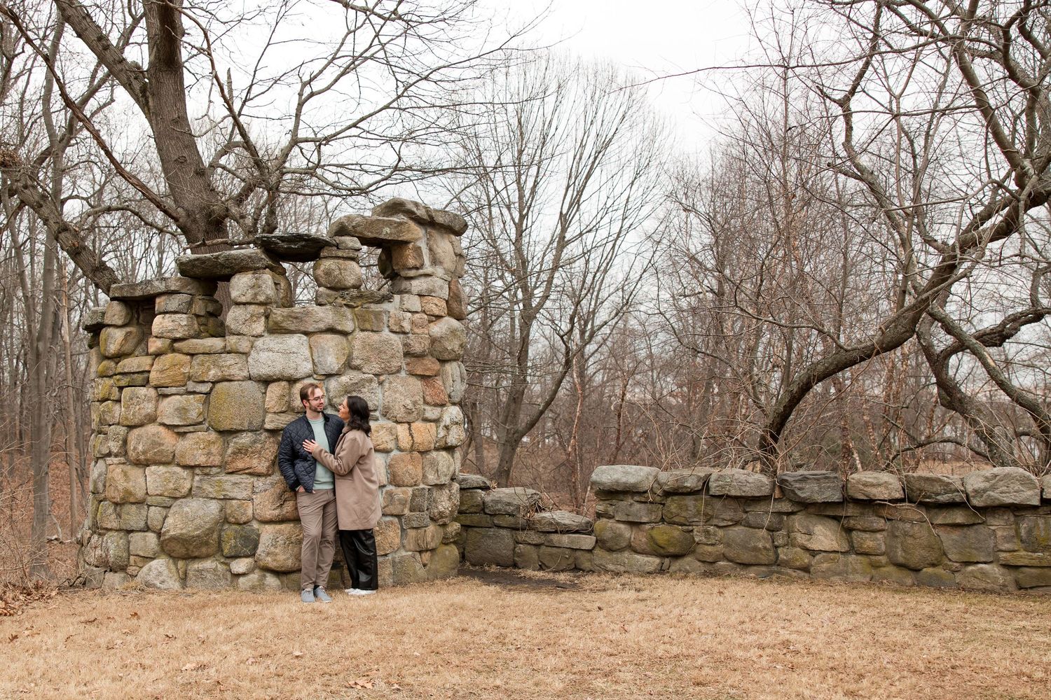 A couple standing together near an old stone structure in a wooded area.