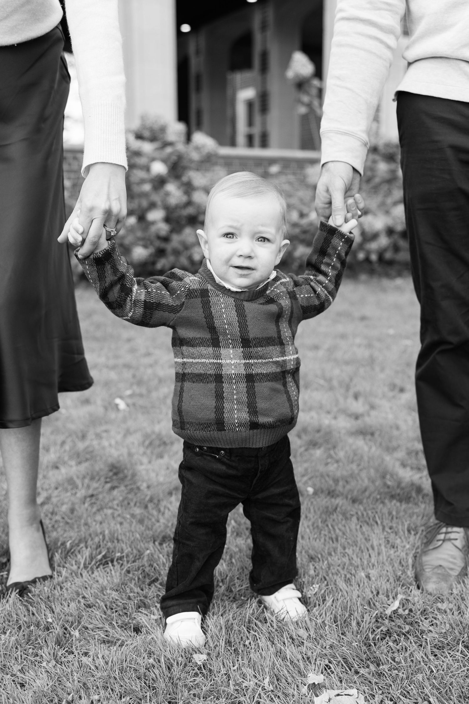 Young child in plaid sweater smiles while standing and holding hands with two adults in black and white photo on lawn.