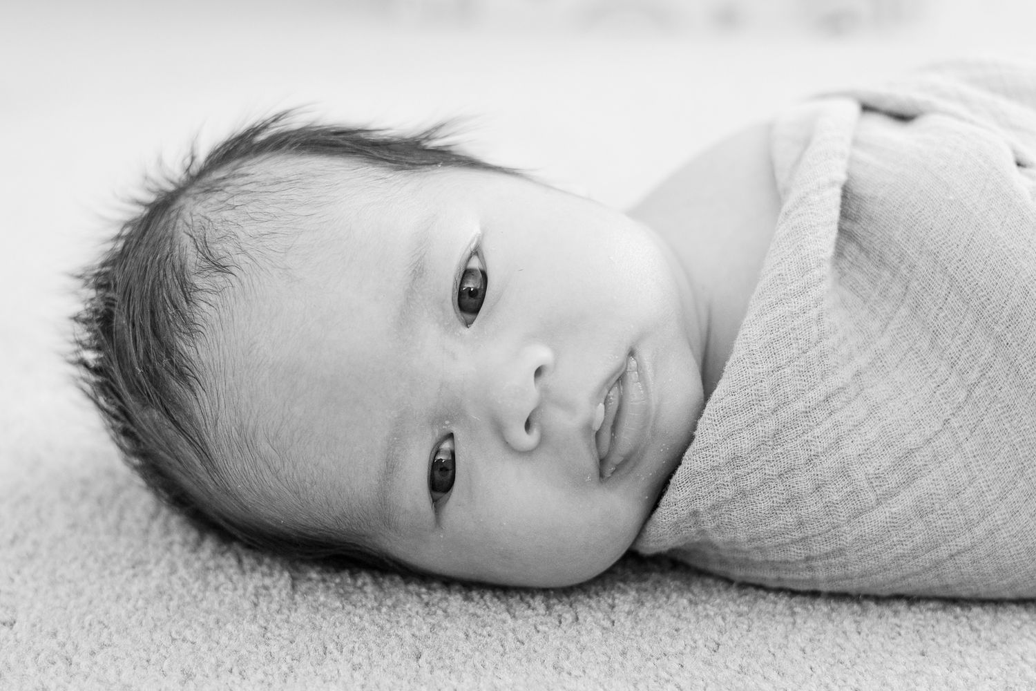 Close-up black and white portrait of an alert newborn wrapped in light textile.