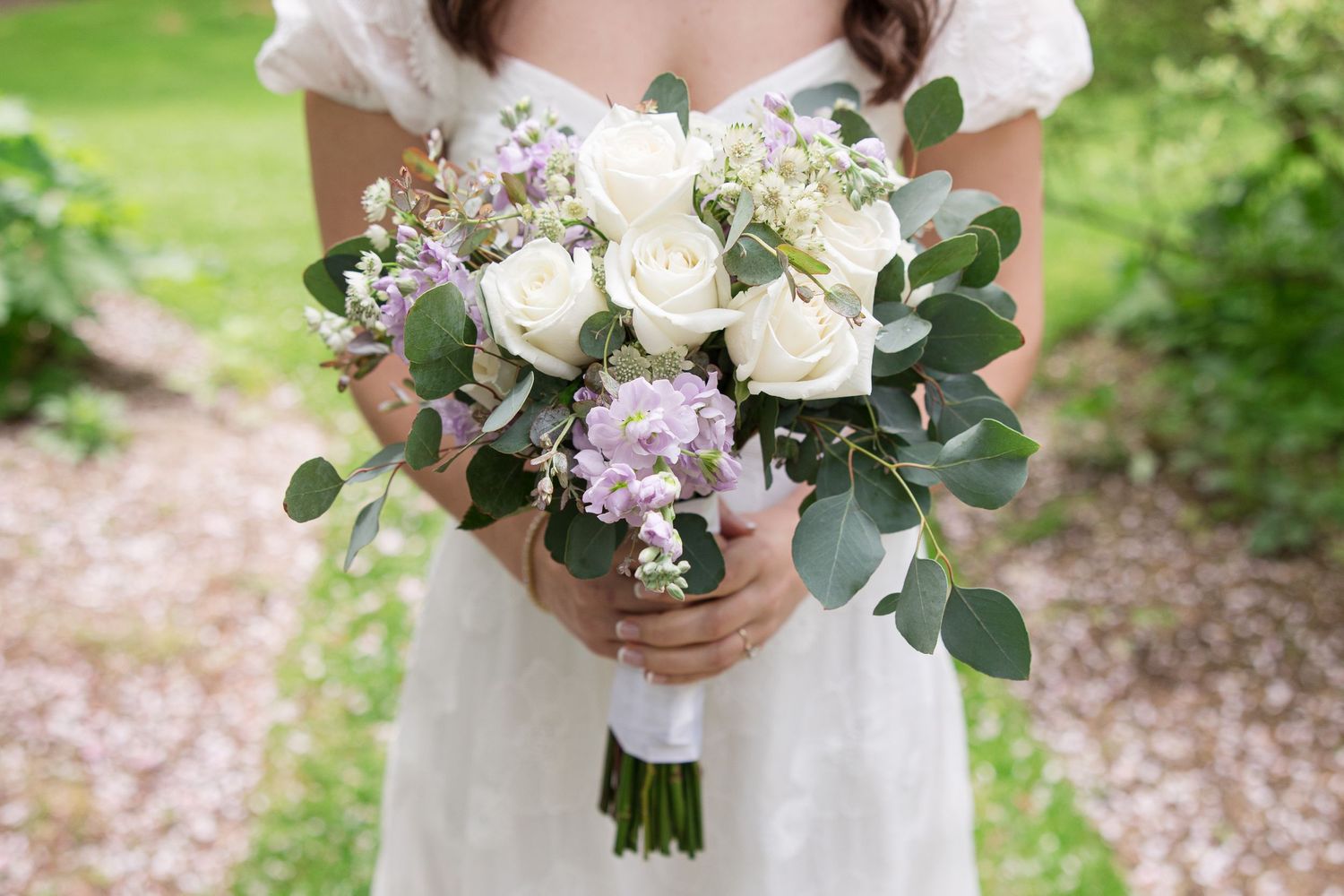 A white and lavender bridal bouquet with roses and eucalyptus leaves held against a wedding dress.