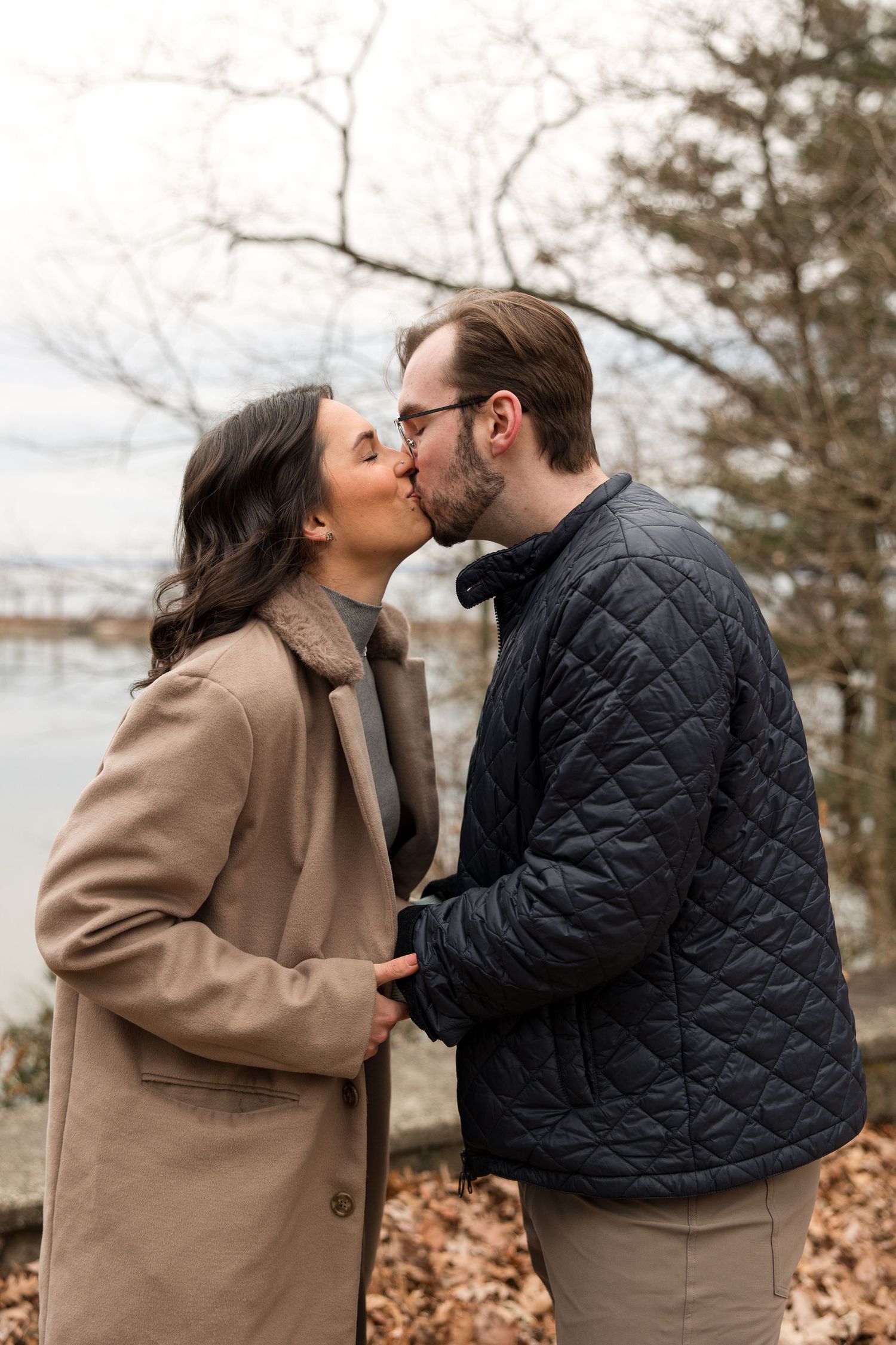 A romantic kiss shared between two people in winter coats by the water.