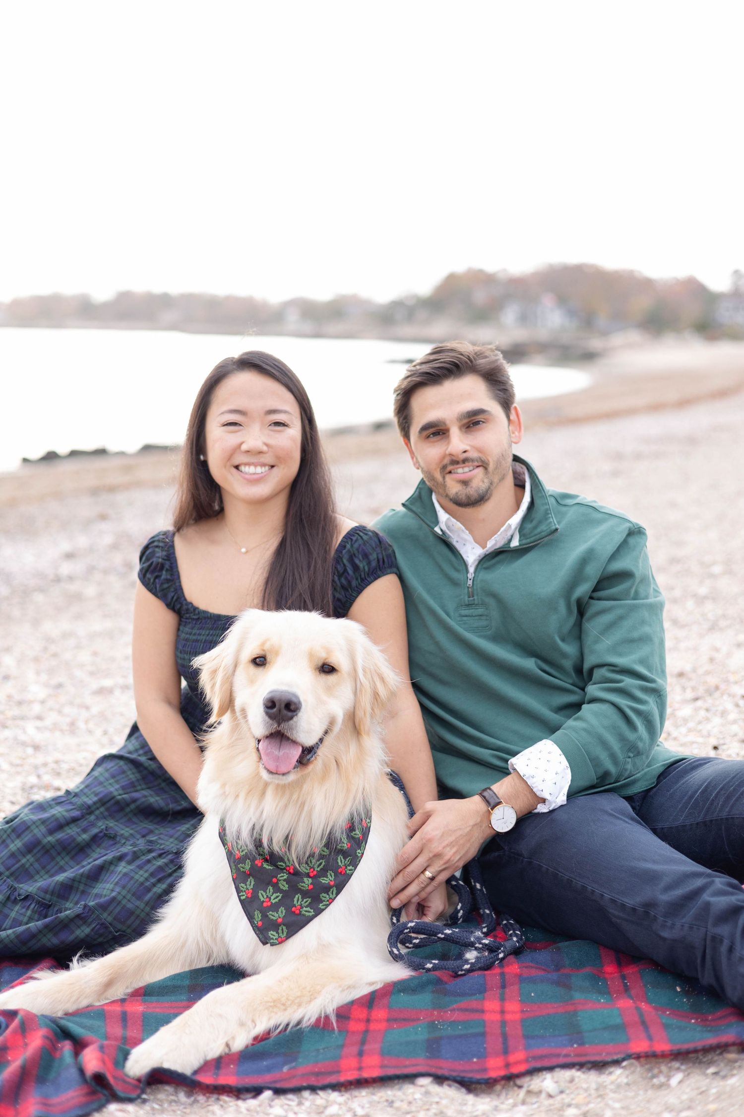 Couple relaxes with their golden retriever on a plaid blanket at the beach during golden hour.