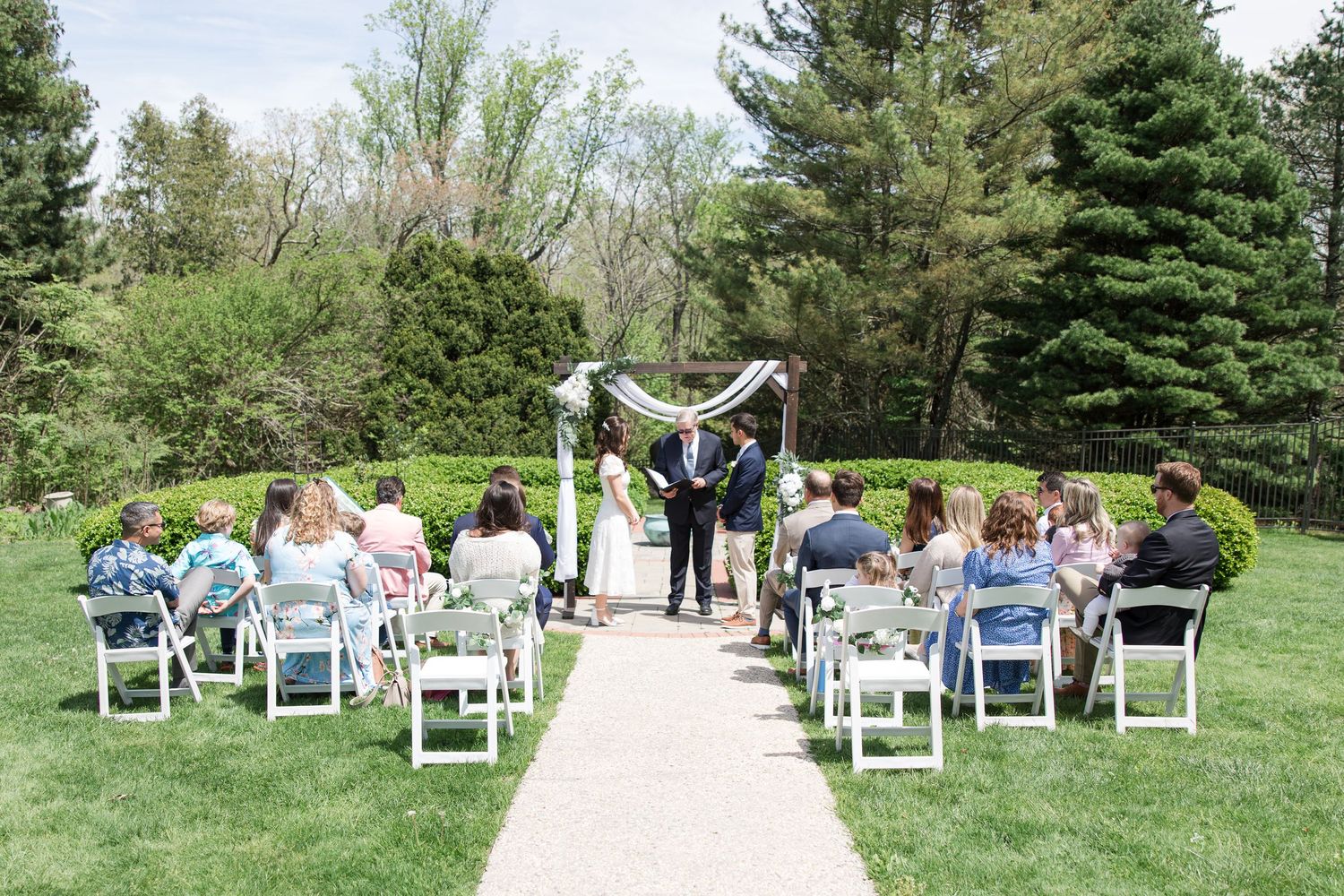 Intimate outdoor wedding ceremony with white chairs arranged on a garden path surrounded by evergreen trees.