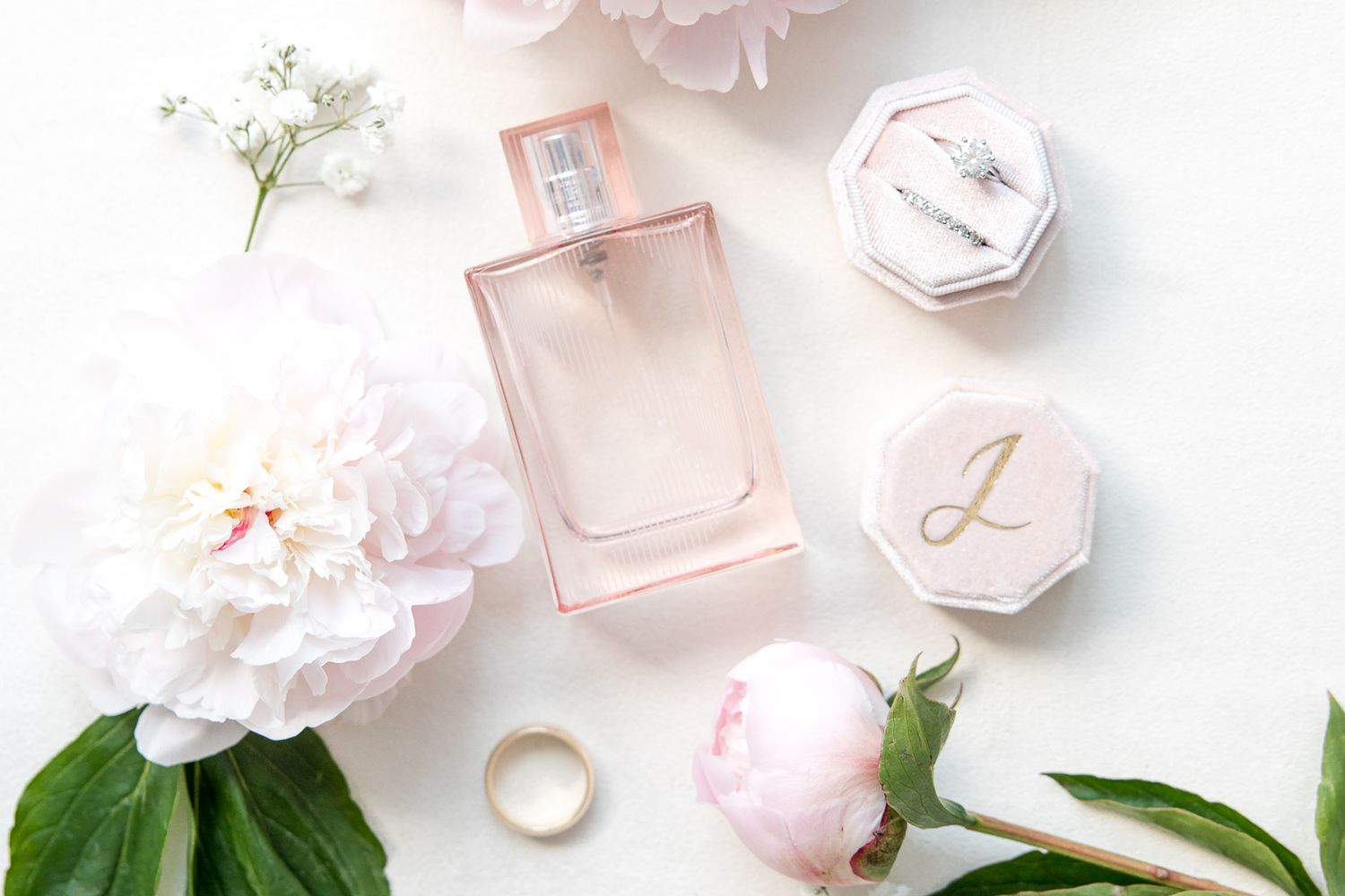 Wedding details styled with pink perfume, flowers, and monogrammed boxes on white background.