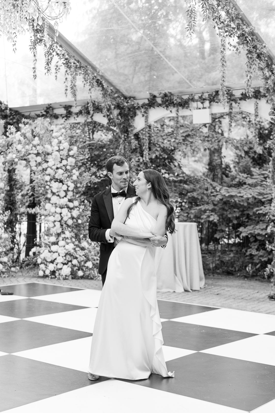 Couple dancing on a black and white checkered floor under a tent decorated with cascading flowers.