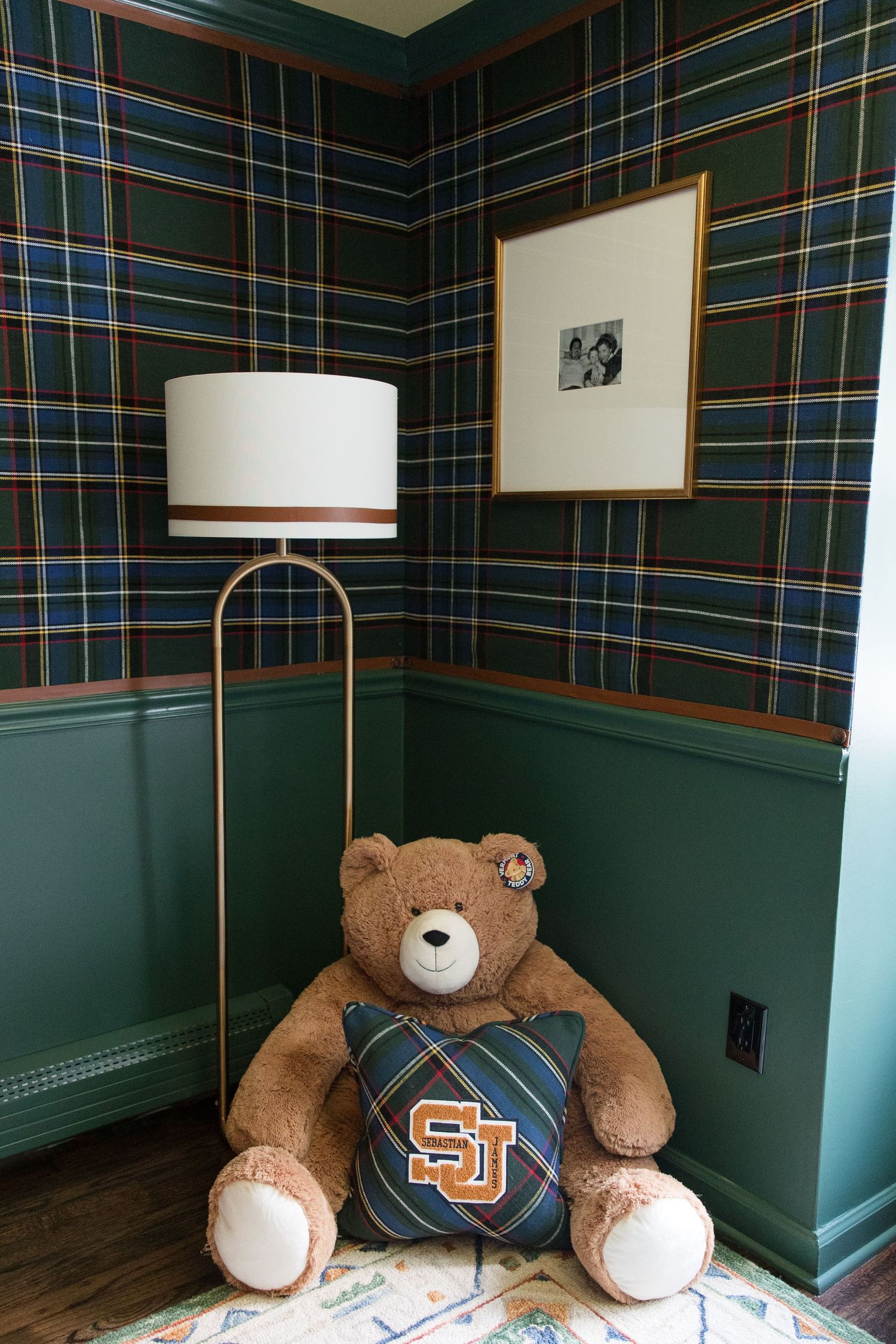 A brown teddy bear wearing an SJ logo sweater sits beneath a modern floor lamp against plaid walls.
