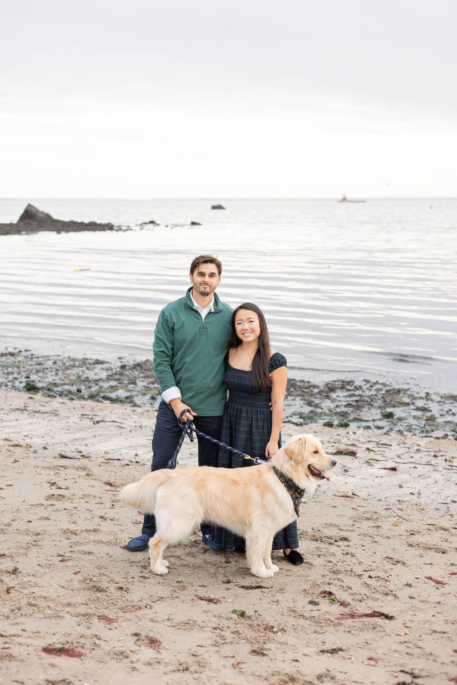 Couple with Golden Retriever dog poses on rocky beach with ocean waves in background.