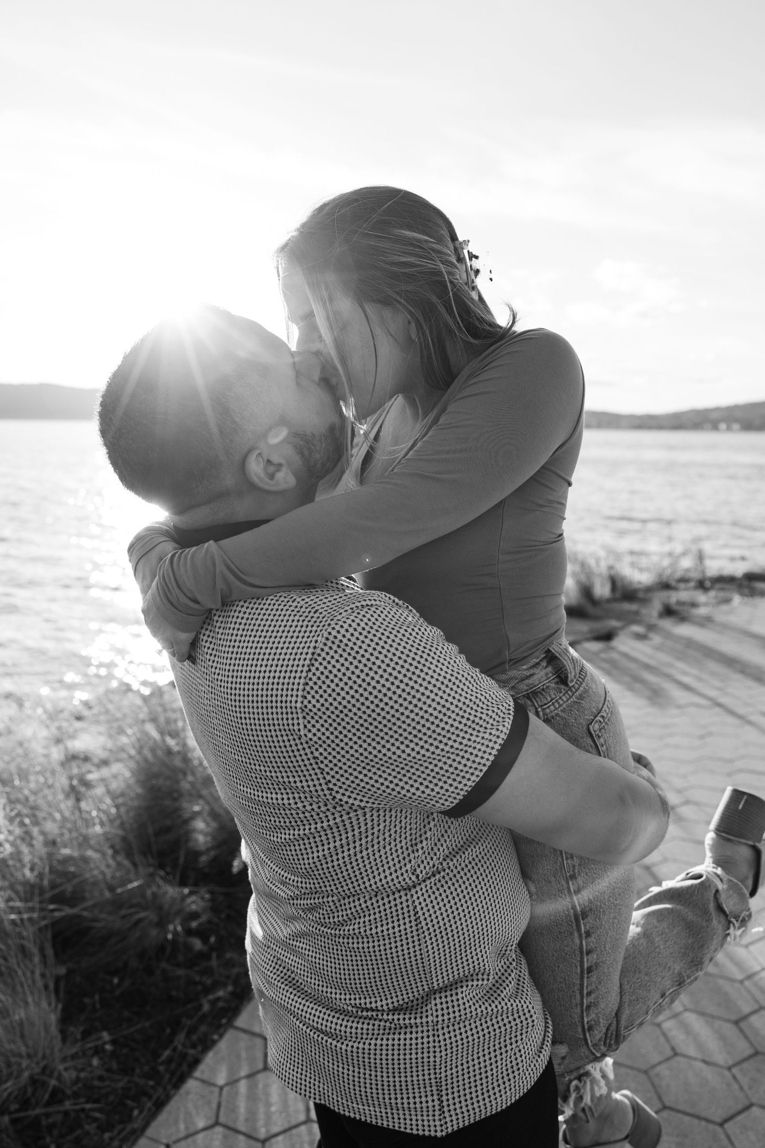 Black and white photo of romantic couple embracing by waterfront at sunset.