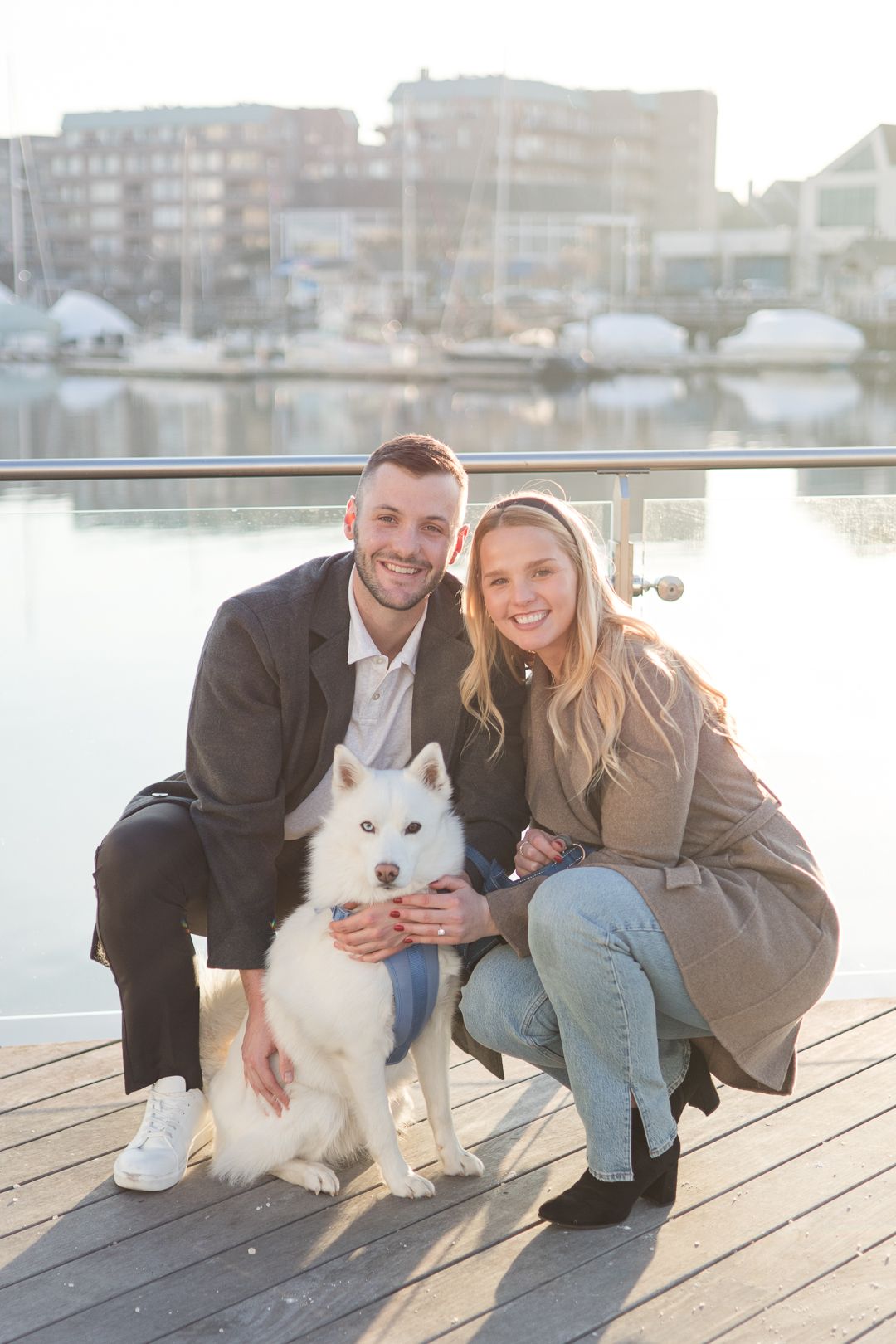 A couple and their white Husky dog pose together on a wooden dock by a marina.