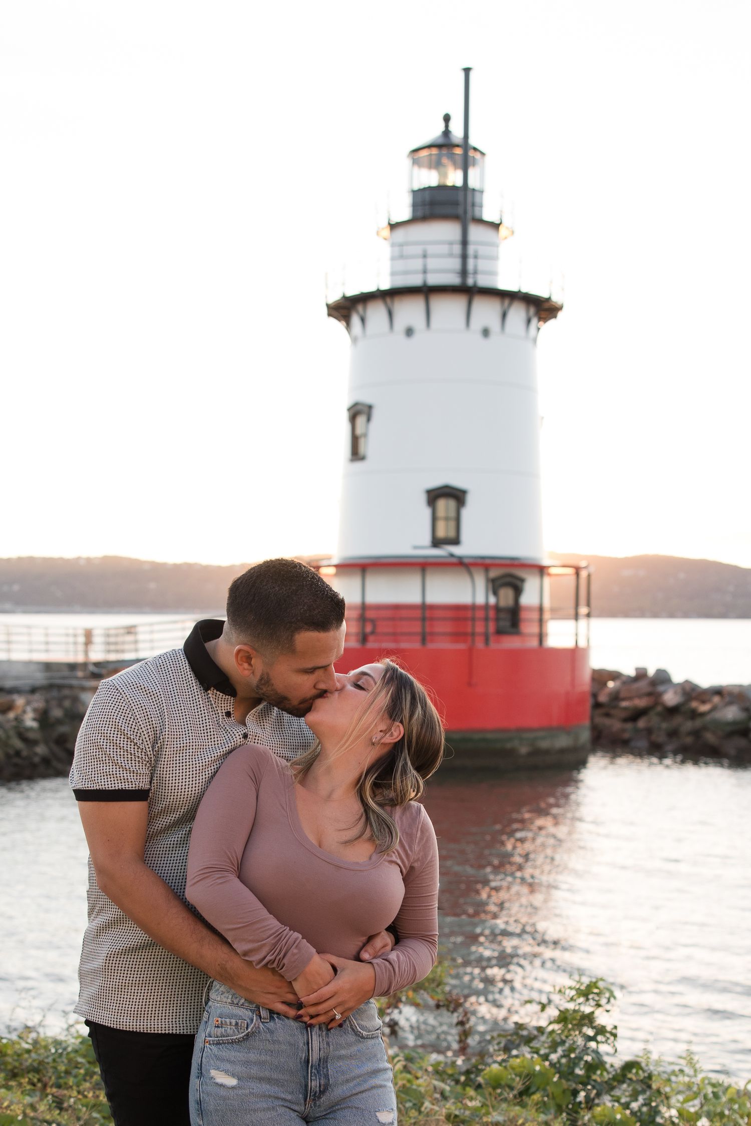 Couple embraces in romantic moment with white and red lighthouse as backdrop.