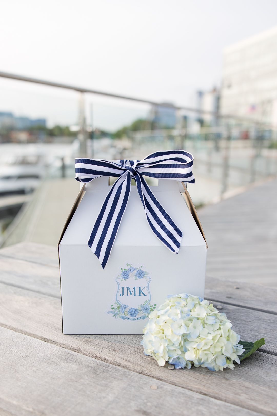 White gift box with navy striped bow and hydrangea flower on wooden dock.