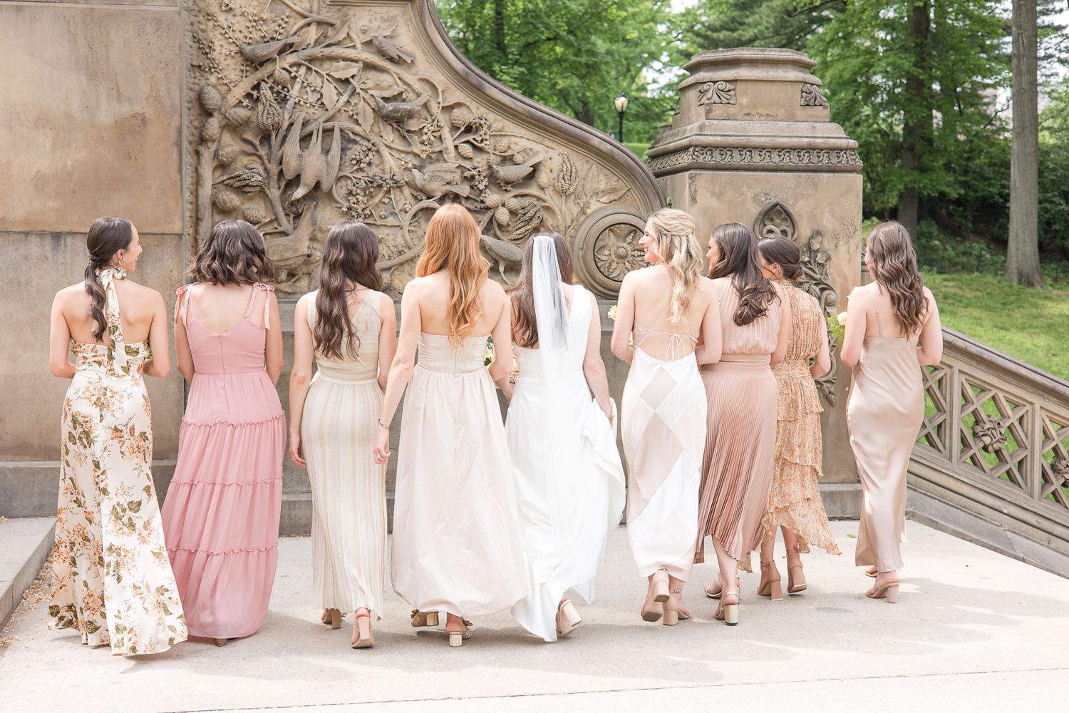 Bridal party walking away showing backs of blush and floral dresses against decorative stone architecture.