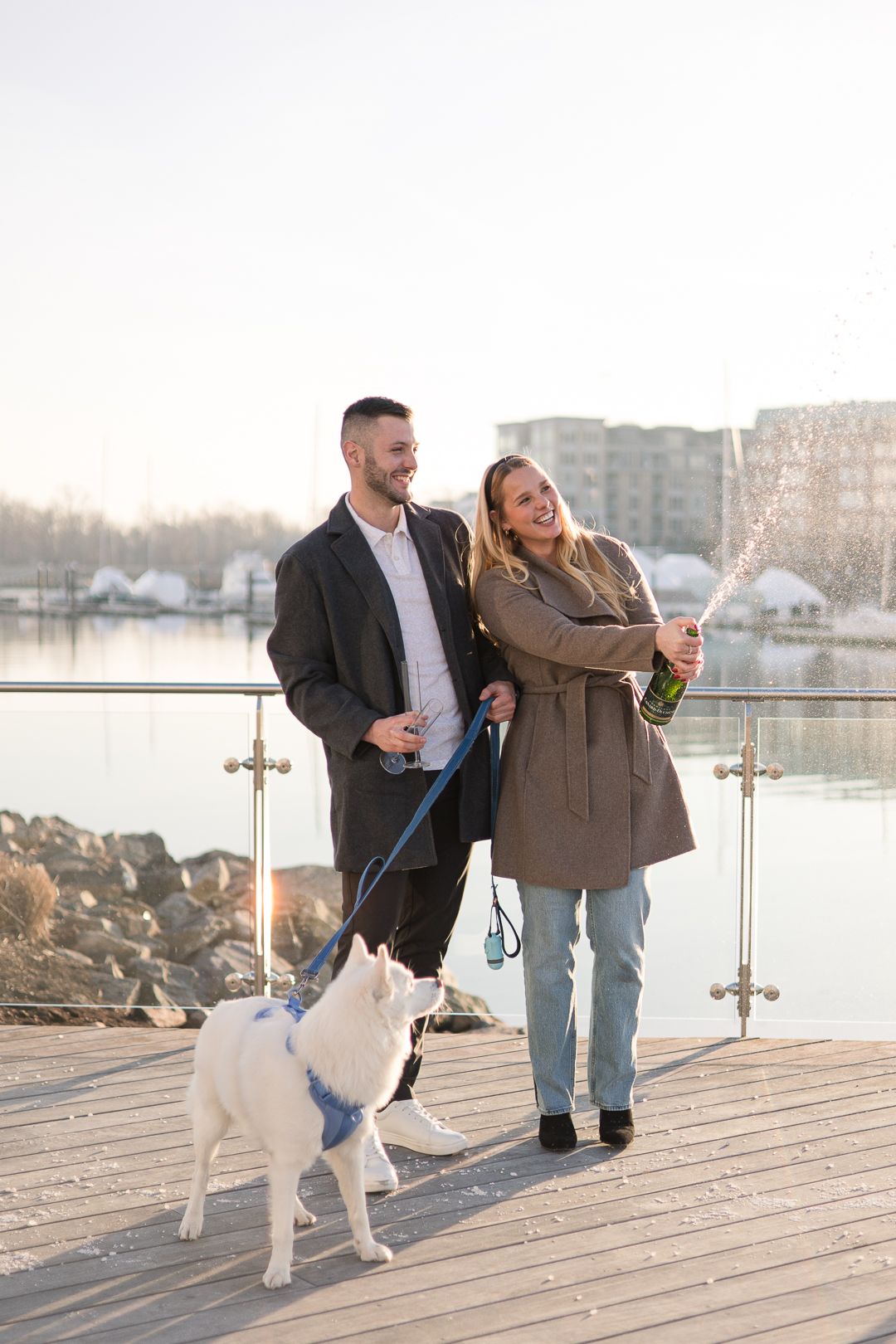 Couple laughing together on marina boardwalk with their white dog during sunset.