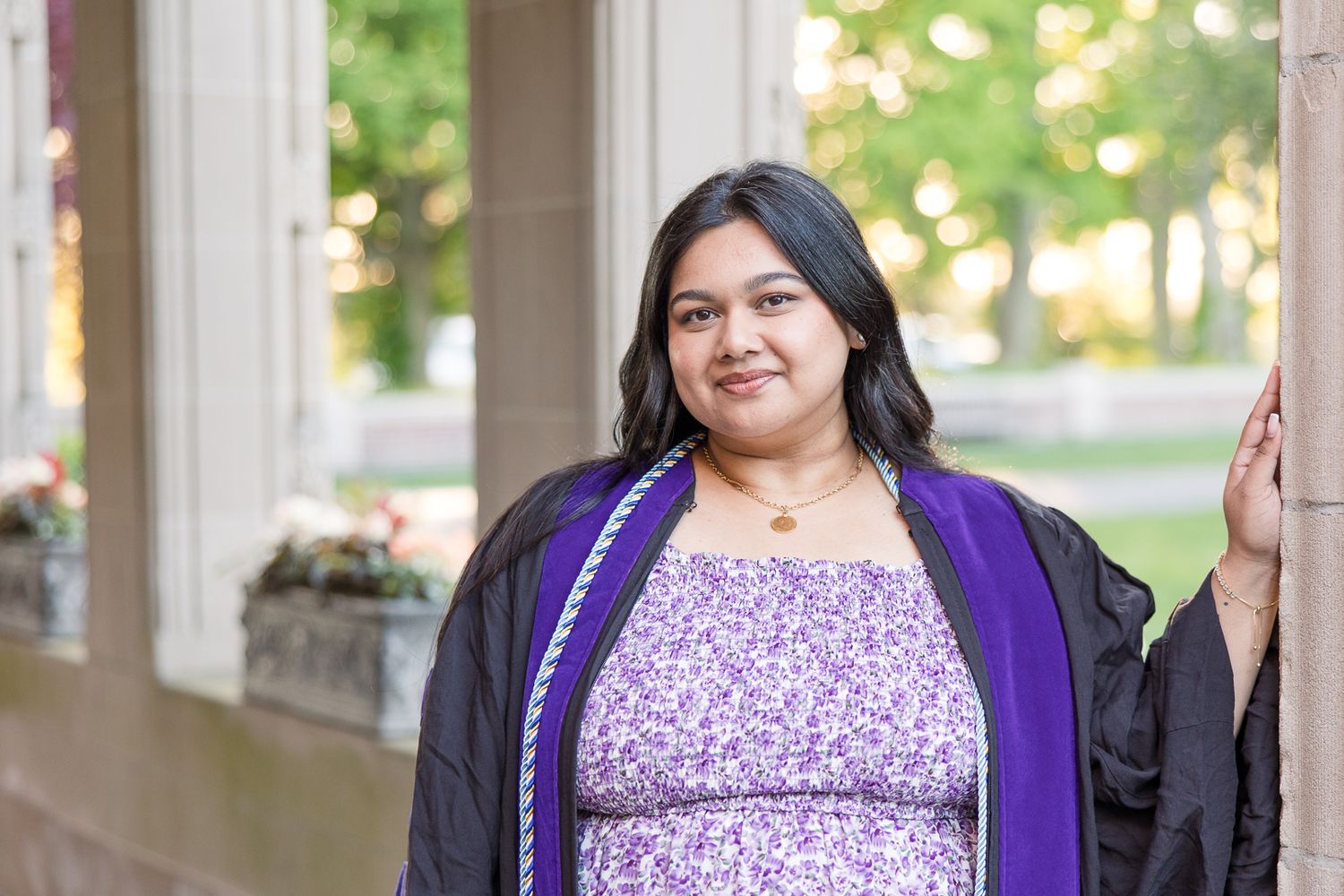Graduate wearing purple academic regalia smiles in outdoor portrait setting.