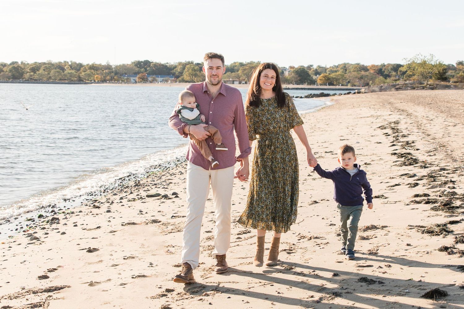 Family strolling together along peaceful beach with water and coastline in background.
