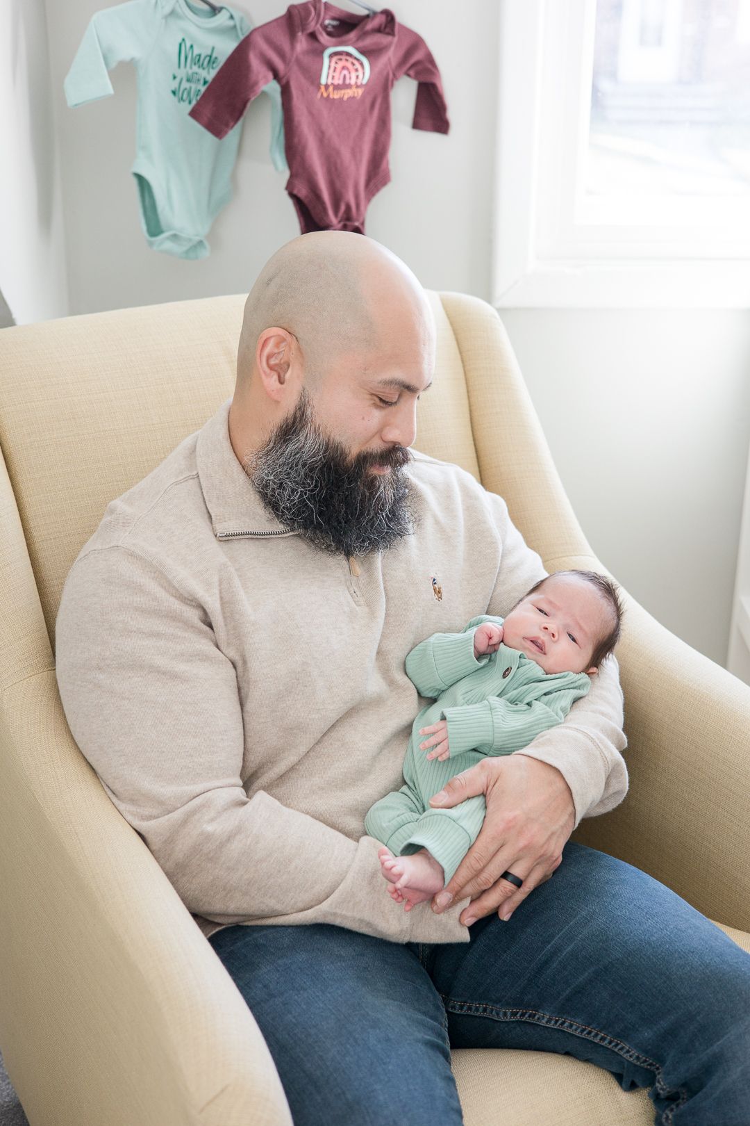 A bearded man in a beige sweater cradles a newborn baby wearing mint green in a cream-colored armchair.