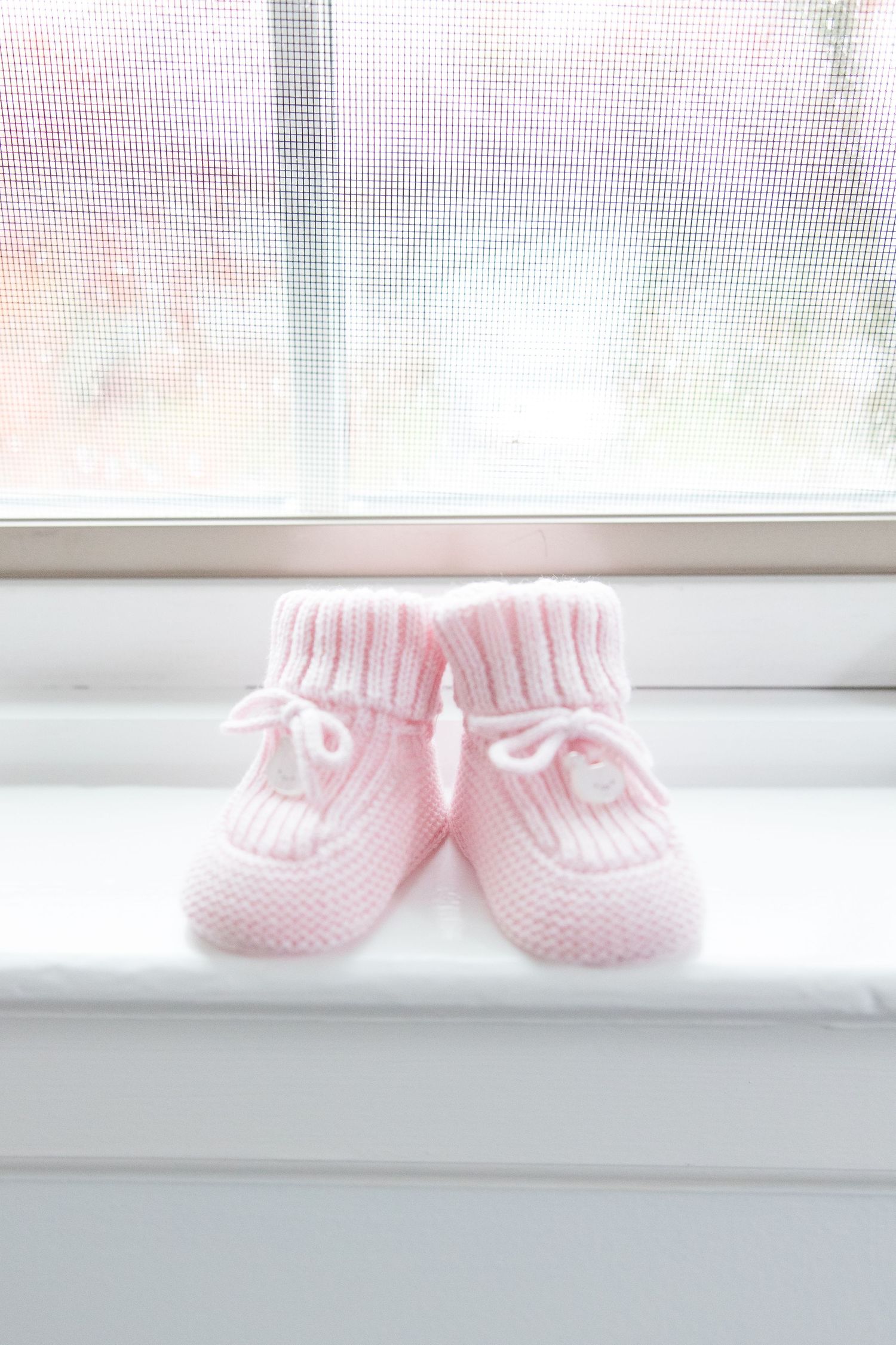 Tiny pink knit baby booties sitting on white windowsill in soft light.