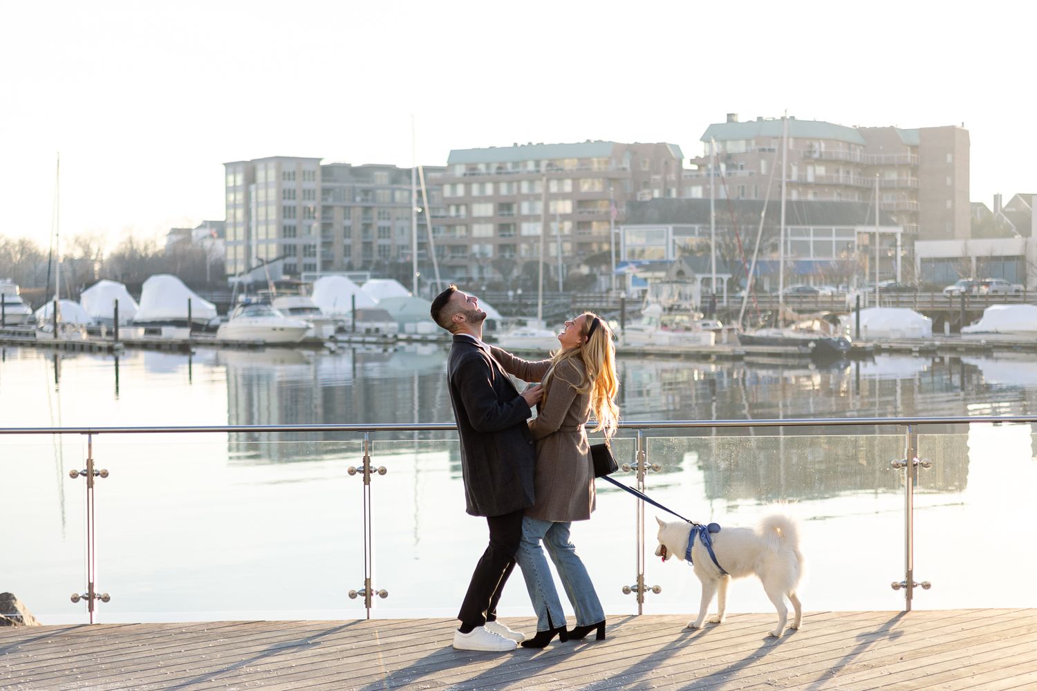 Happy couple dancing on a waterfront deck with their white dog during sunset.