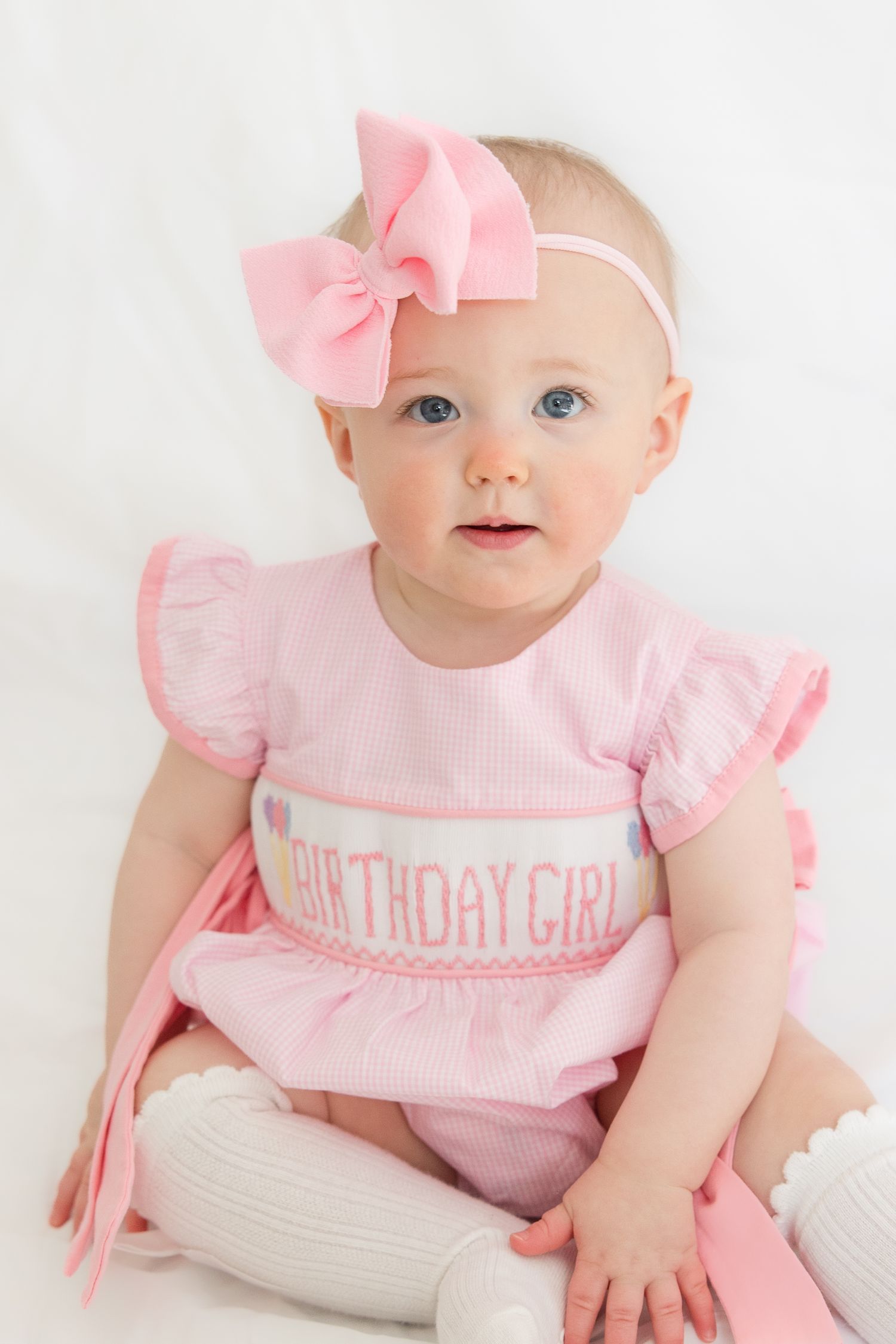 Baby wearing pink birthday dress with matching bow headband and white knee socks sits on white background.