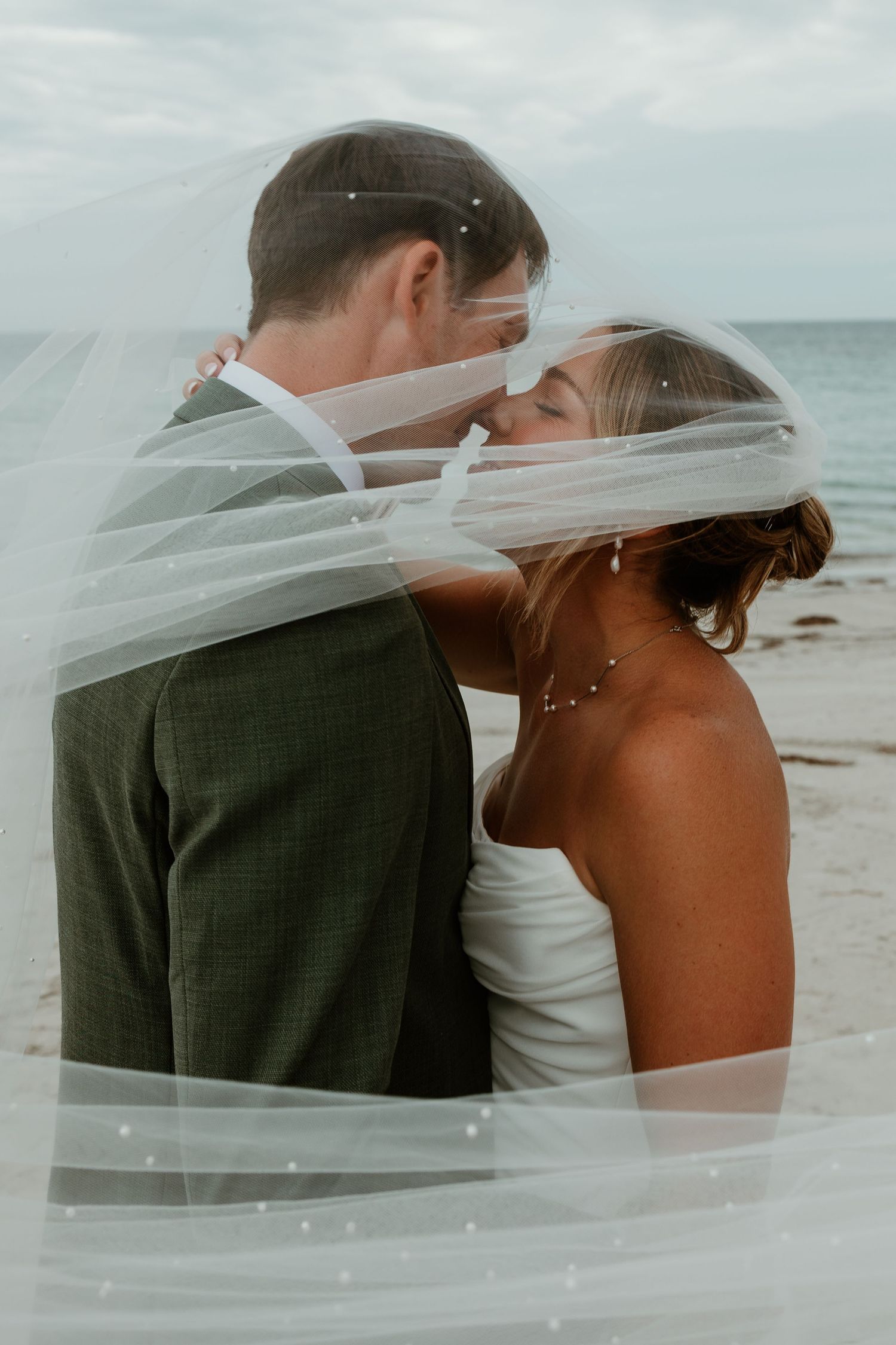 Bride and groom sharing a romantic kiss on the beach, draped under a flowing wedding veil by the ocean.