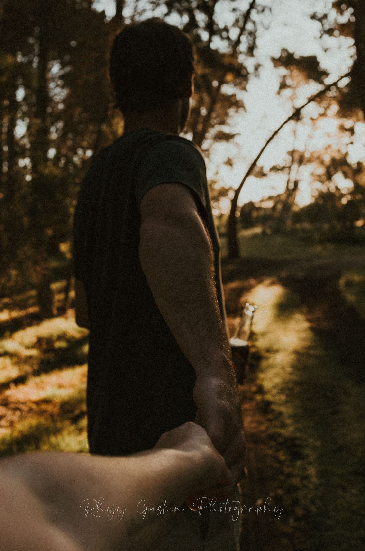 A silhouetted figure holding someone's hand leads the way through a golden sunset-lit forest path.