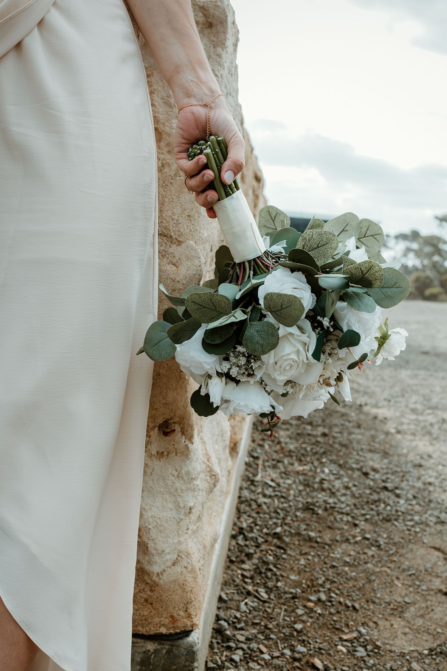 Bride in white dress holding elegant bouquet of white roses and eucalyptus greenery outdoors near stone wall.