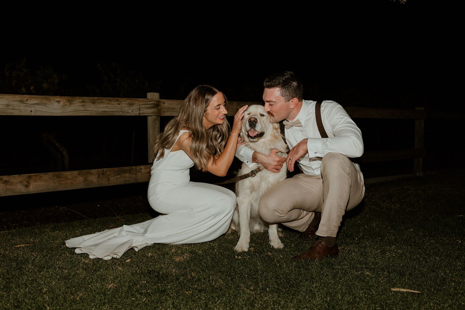 Couple in wedding attire kneels on grass at night with their white dog during outdoor celebration.