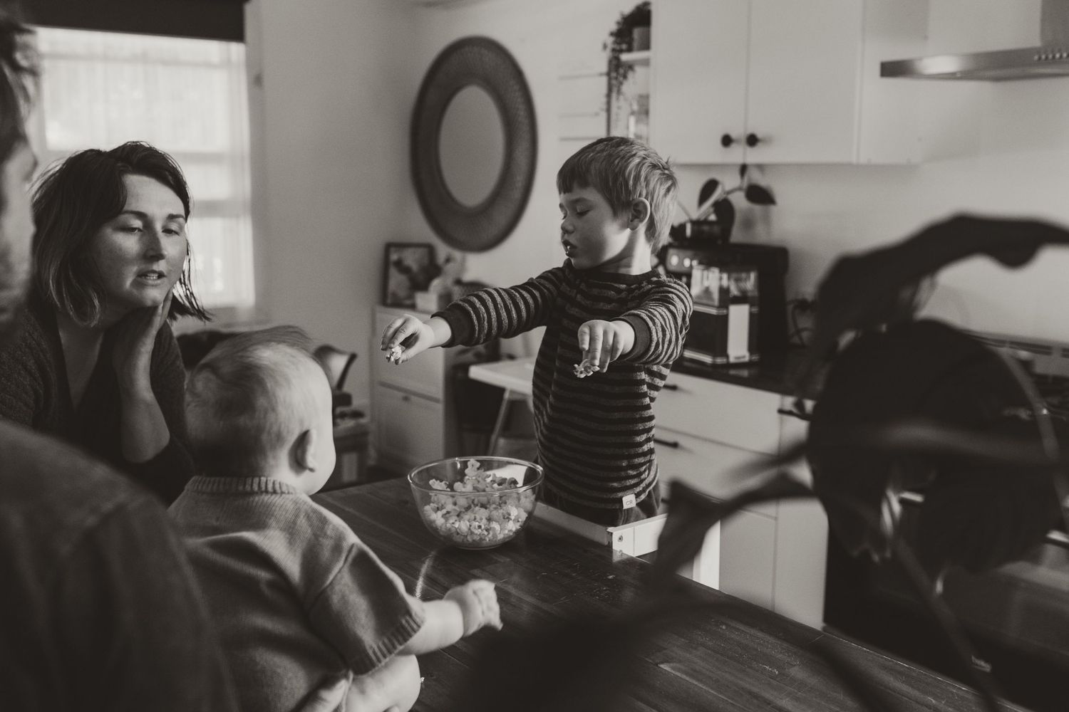 A black and white candid photo of people gathered around a table in a cozy kitchen interior.